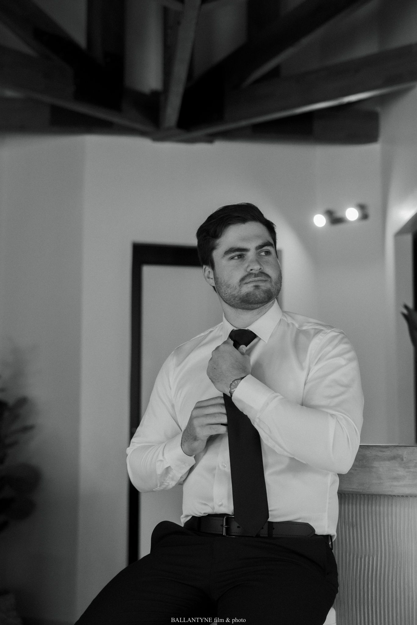 Black and white photo of a groom adjusting his tie while getting ready for his wedding day.