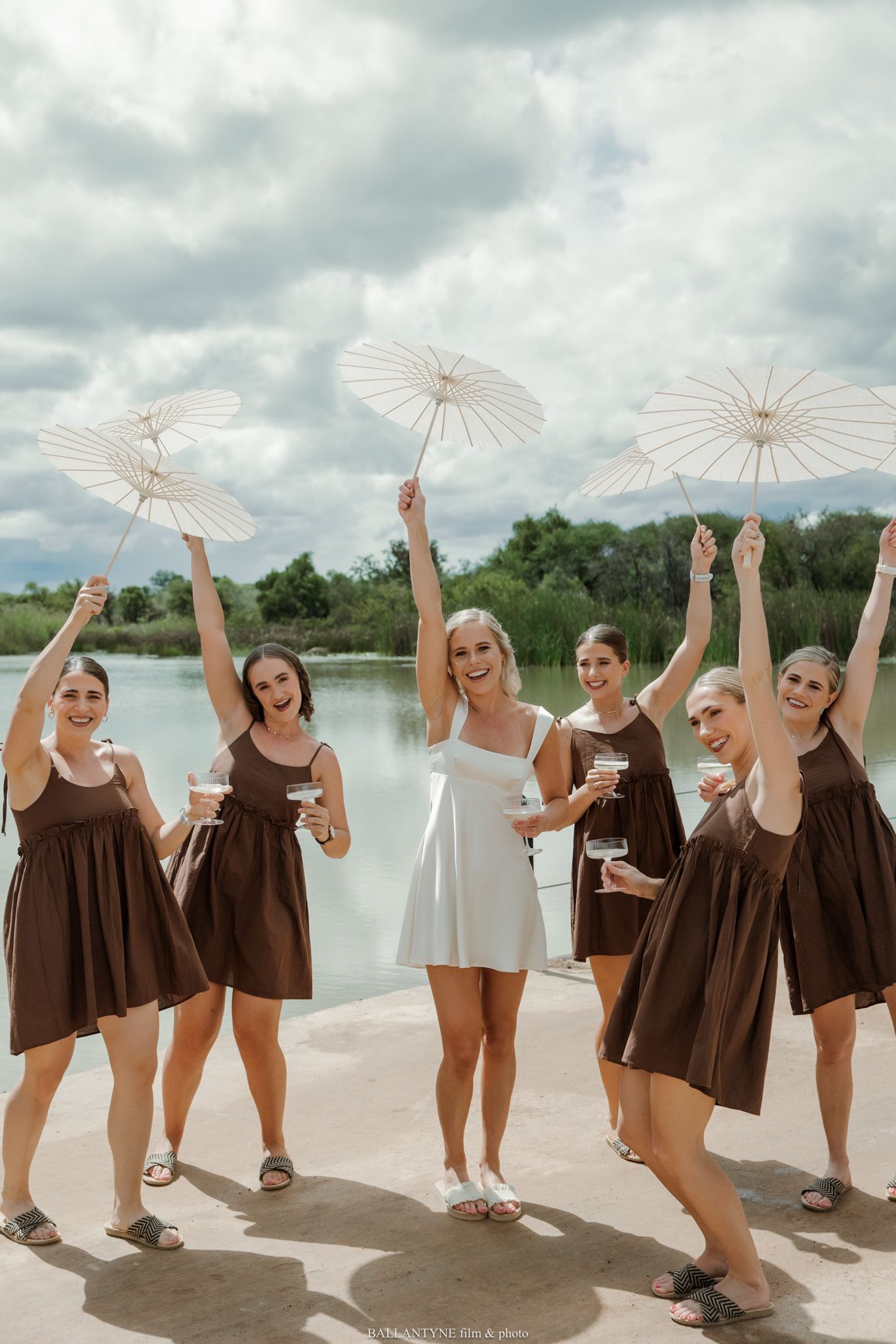 Bride in white dress with bridesmaids in brown dresses holding parasols celebrating outdoors by a lake.