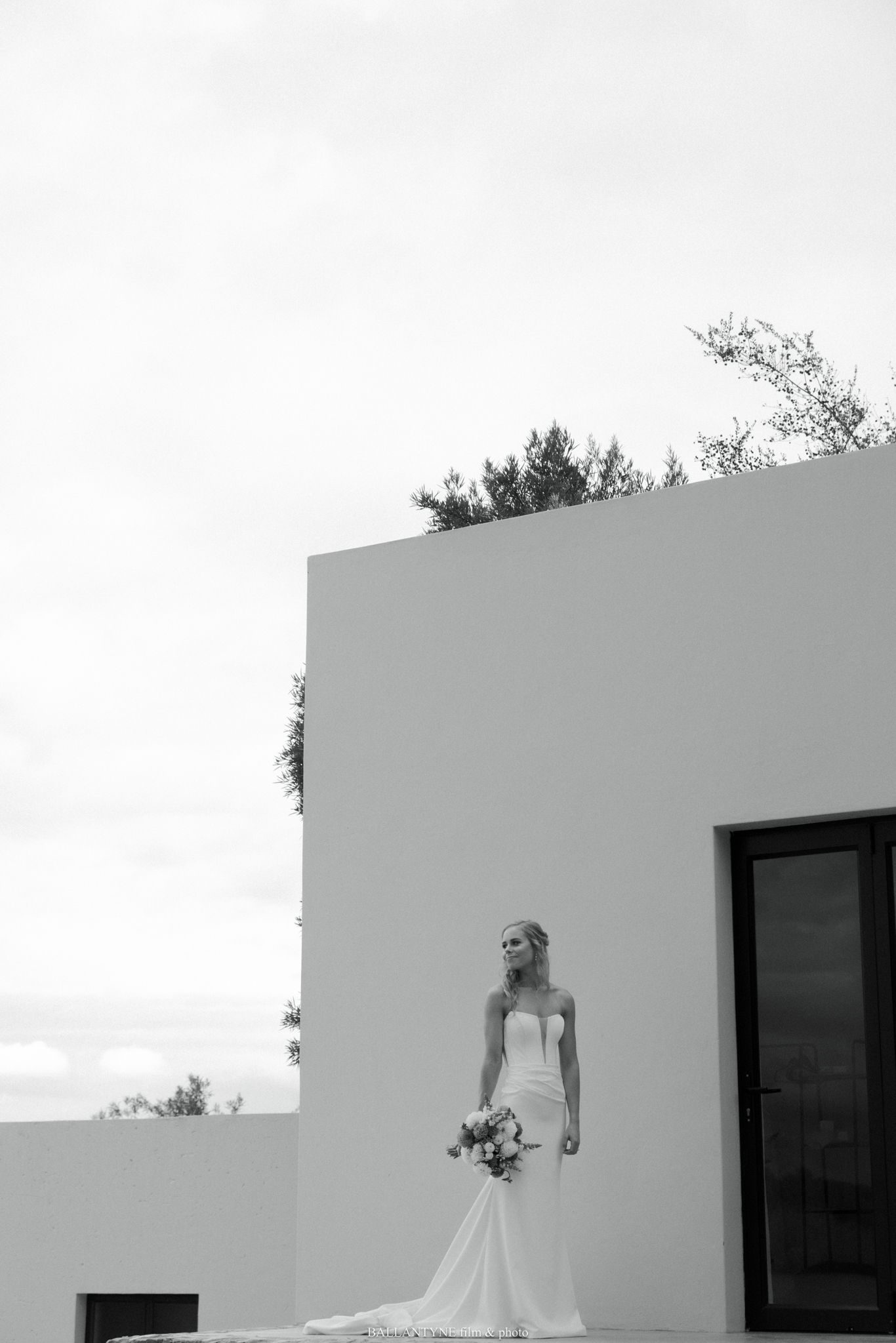 Bride in strapless white gown holding bouquet stands before modern white building in elegant black and white wedding photo.
