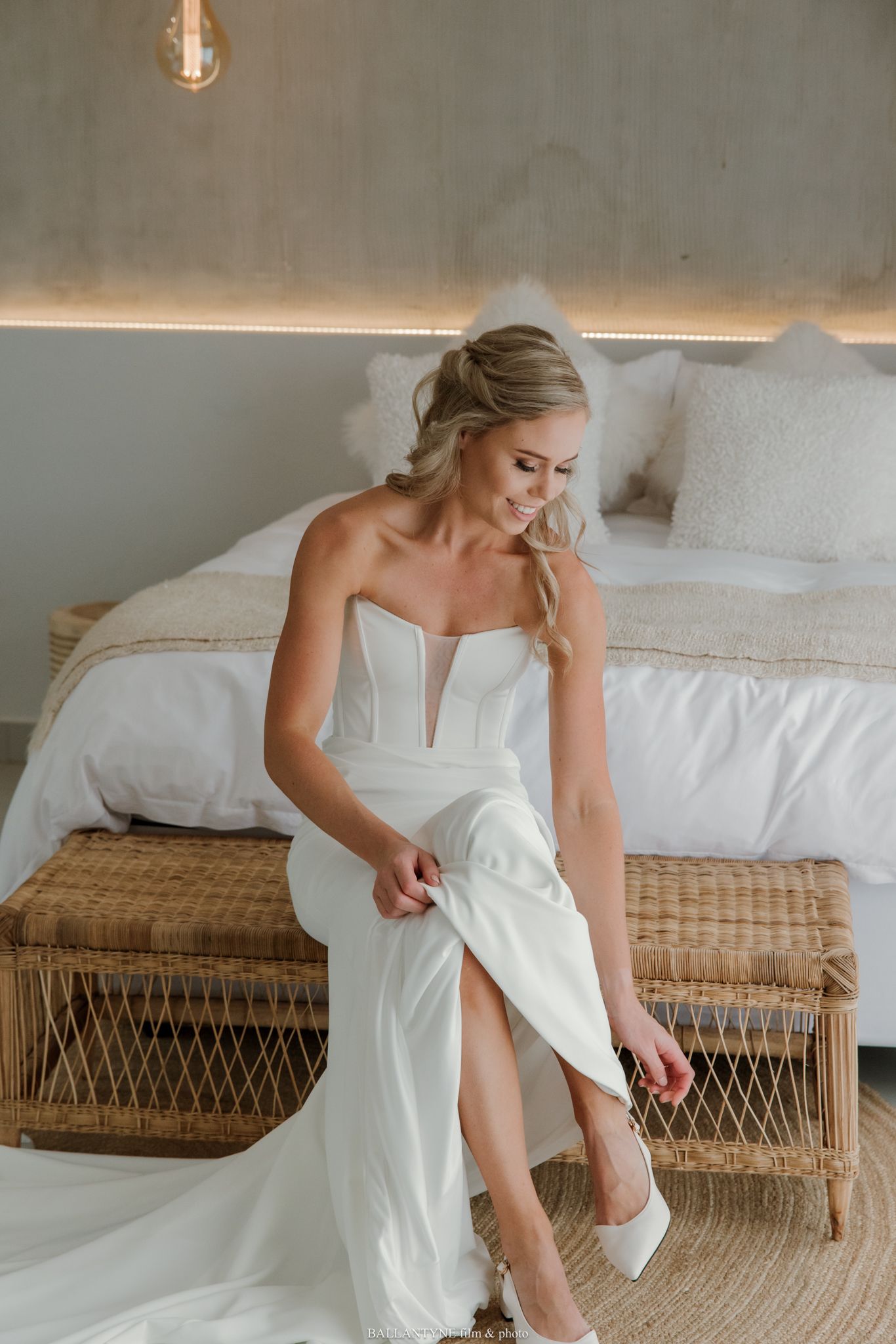 Bride in strapless white wedding gown with plunge neckline sits on wicker bench adjusting her white heels.