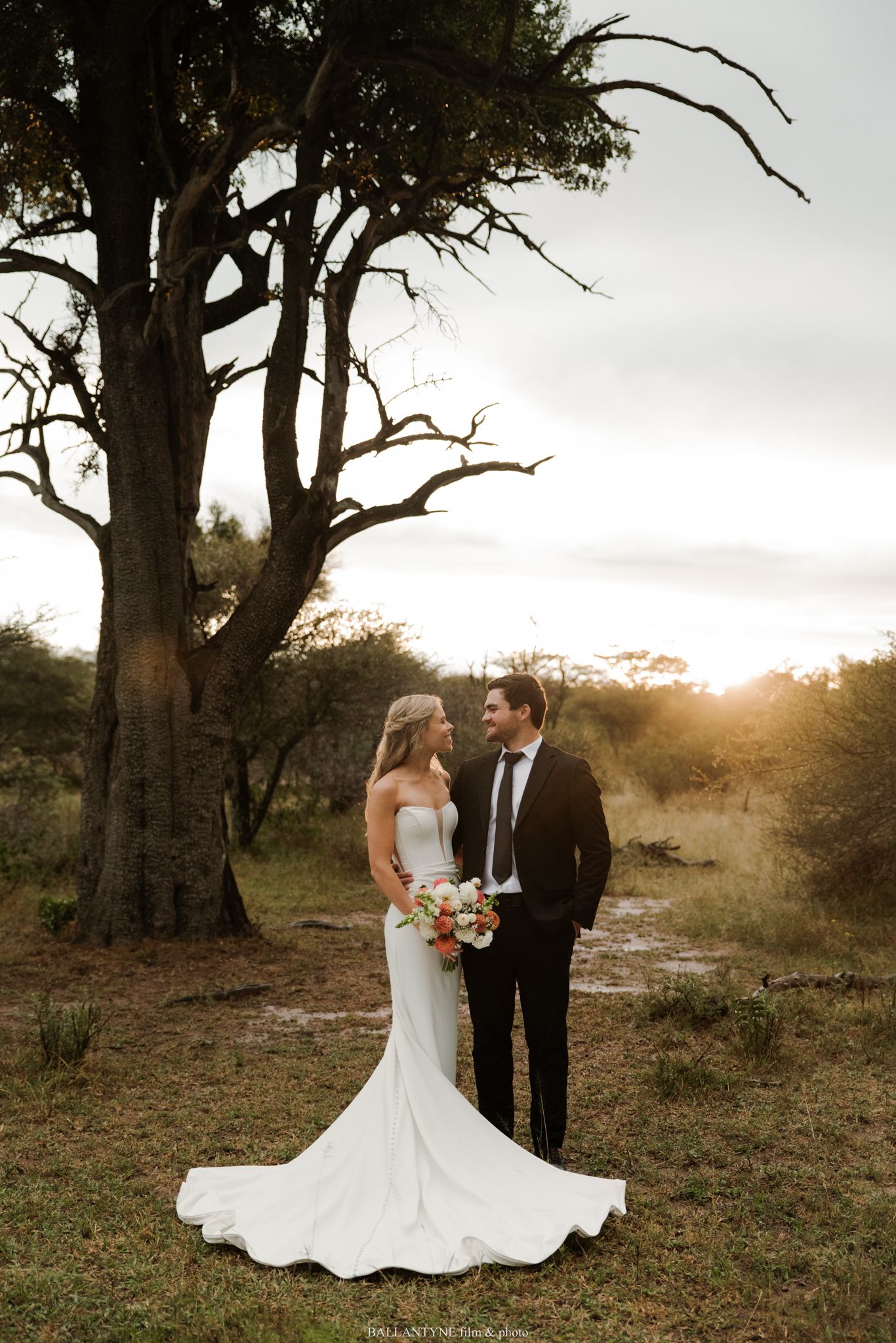 Bride and groom share a romantic moment at golden hour beside a large tree in a South African bush wedding setting.