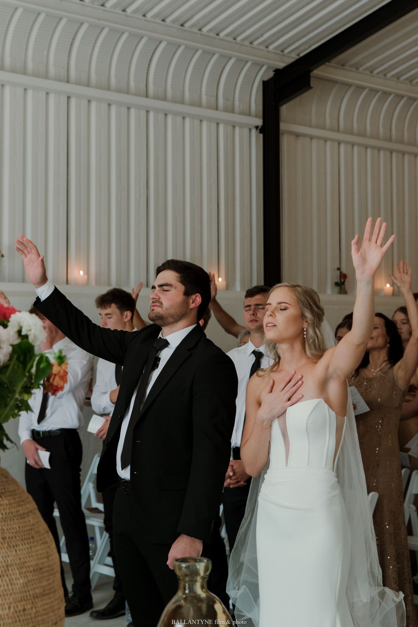 Bride and groom raise their hands in worship during an elegant indoor wedding ceremony with guests standing behind them.