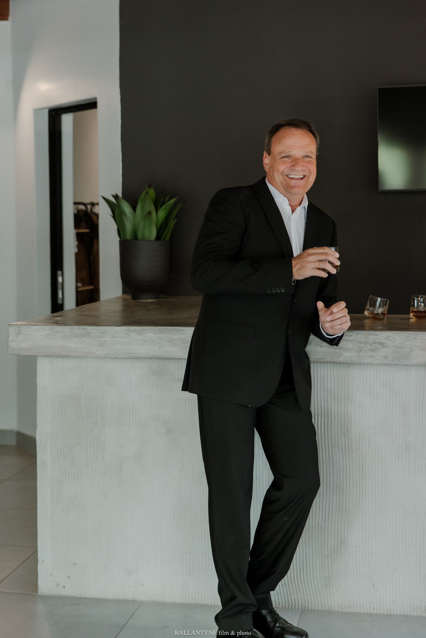 Smiling man in black suit leaning against modern bar counter, holding drink in contemporary interior space.