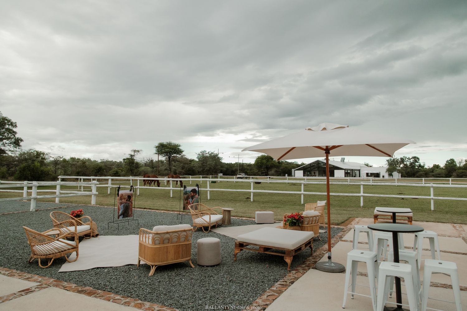 Outdoor lounge area with wicker furniture and umbrella on gravel patio near white fence at rustic farm venue.