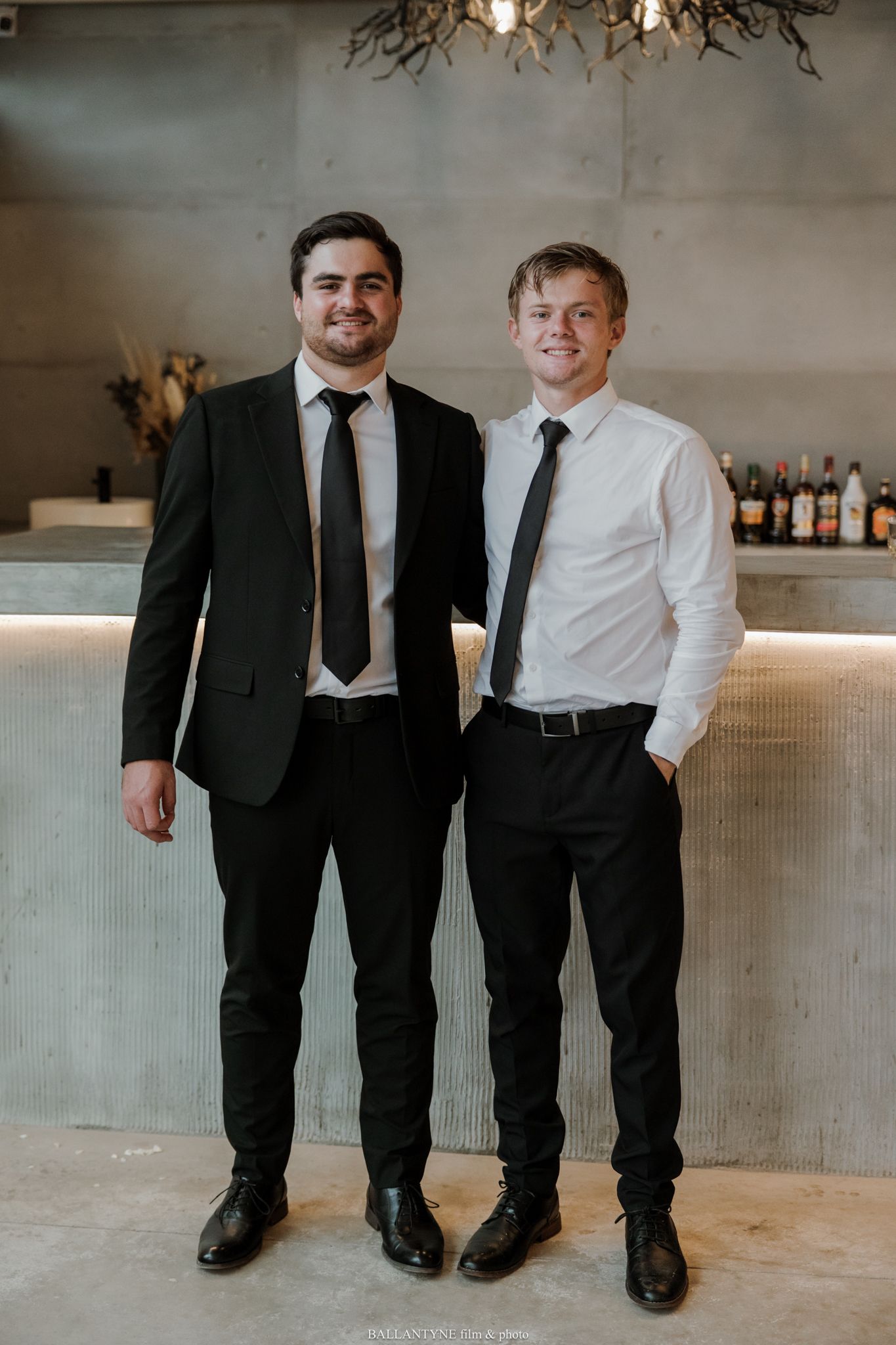 Two men in black trousers and ties pose smiling in front of a bar backdrop.