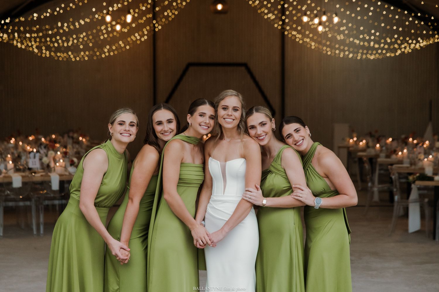 Bride in white gown poses with four bridesmaids in olive green dresses inside a warmly lit barn venue.