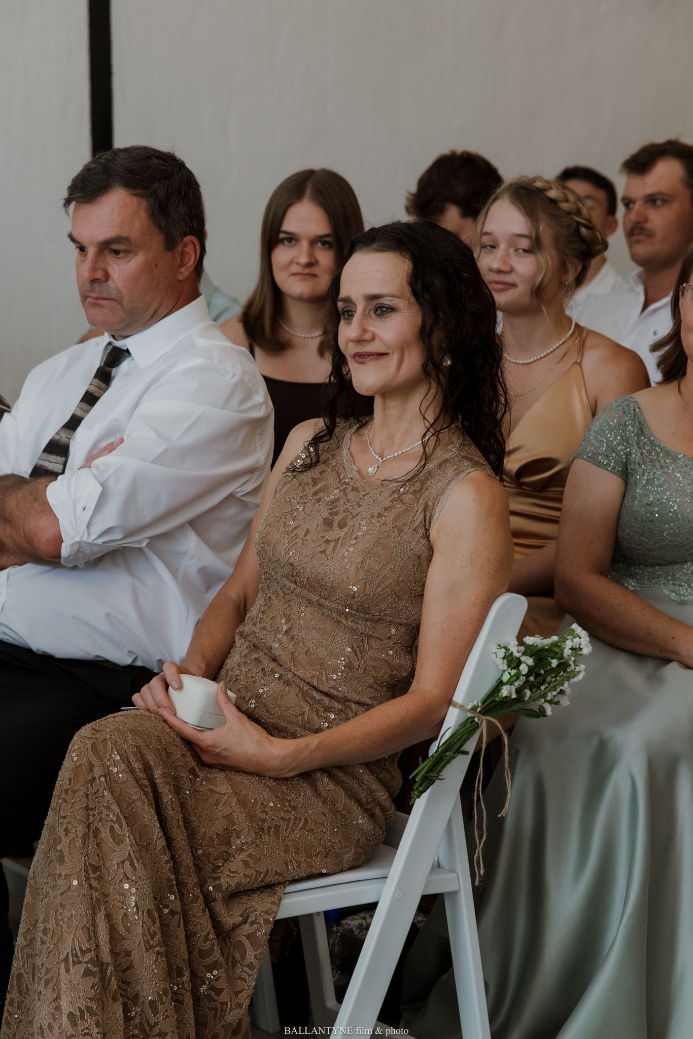 Wedding guests seated at an indoor ceremony, woman in gold lace dress smiling warmly in the foreground.
