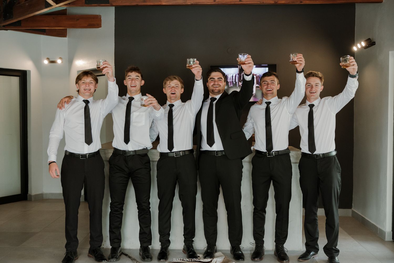 Seven groomsmen in white shirts and black ties raise glasses in a celebratory toast before a wedding ceremony.