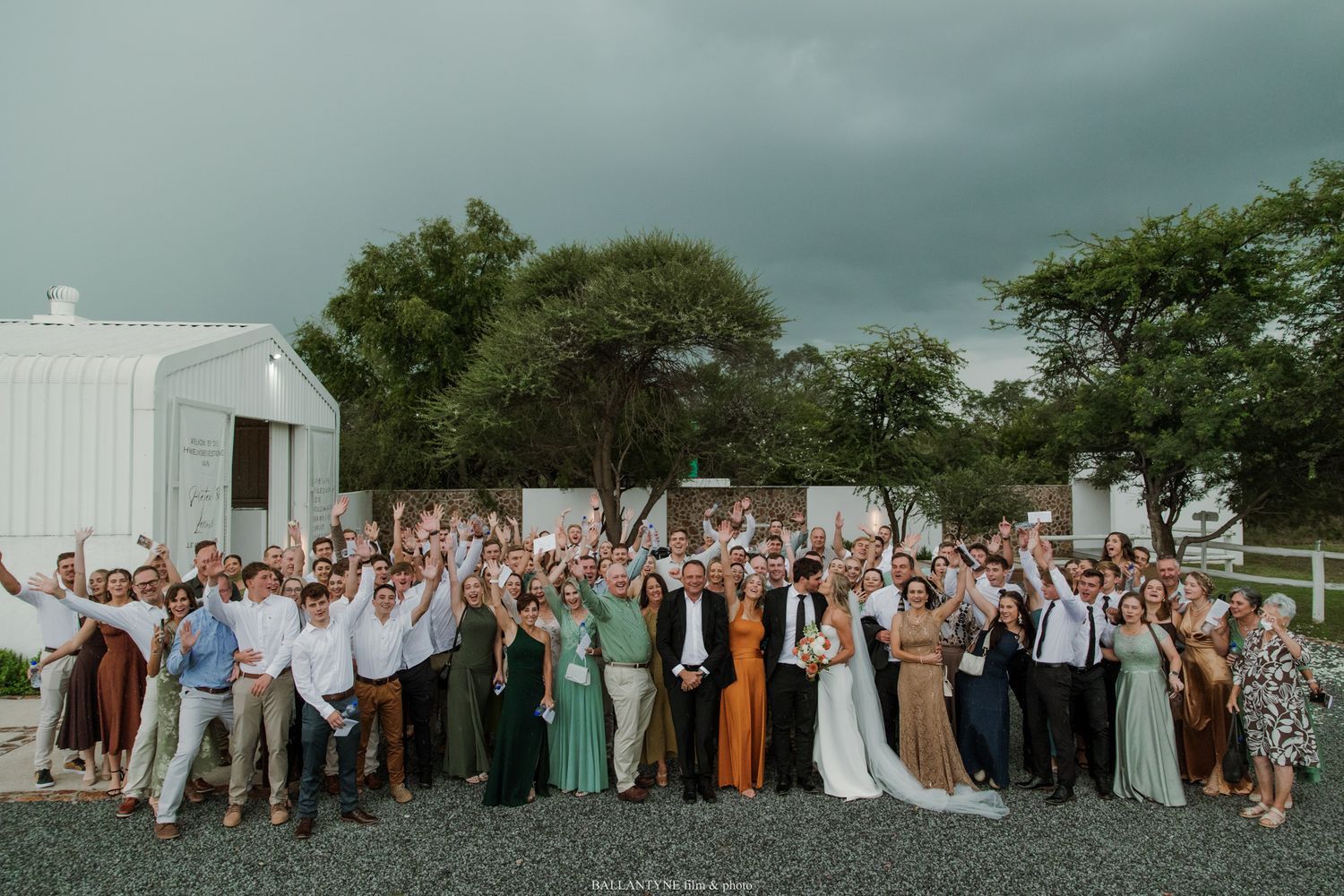 Large wedding group photo with bride and groom surrounded by cheering guests outdoors near a white barn.