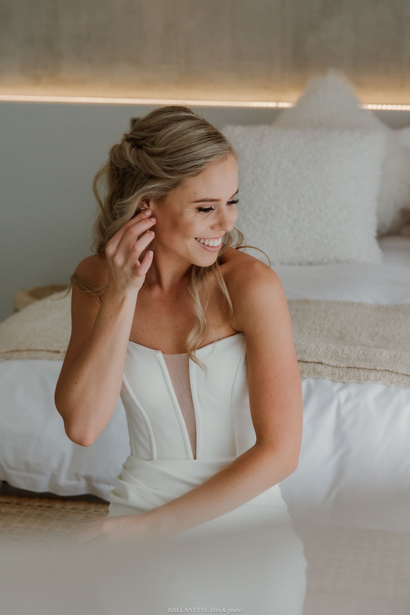 Smiling bride in a white strapless gown adjusting her earring while seated on a bed.