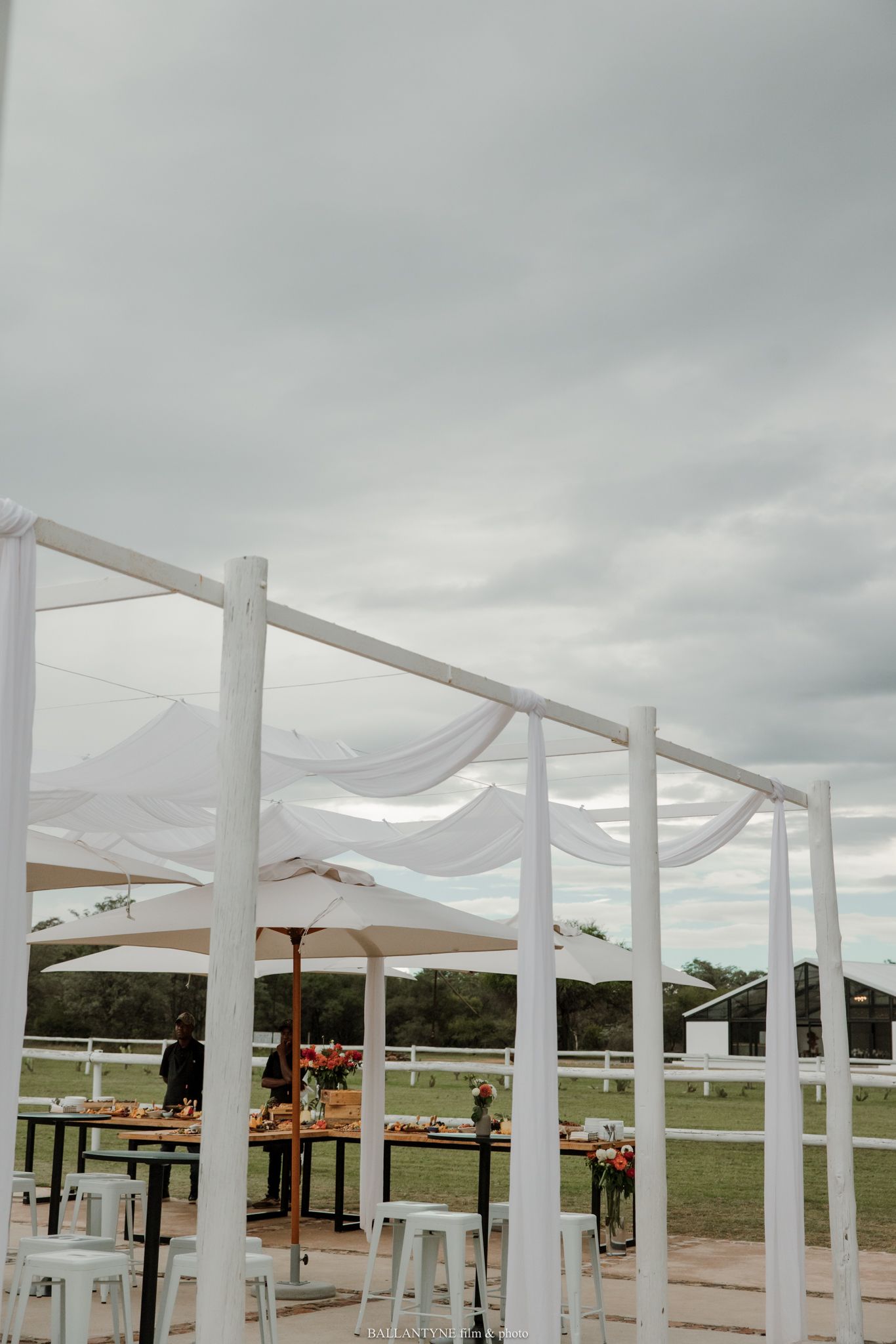 Outdoor wedding reception setup with white draped fabric canopy, tall bar tables, and floral arrangements under a cloudy sky.