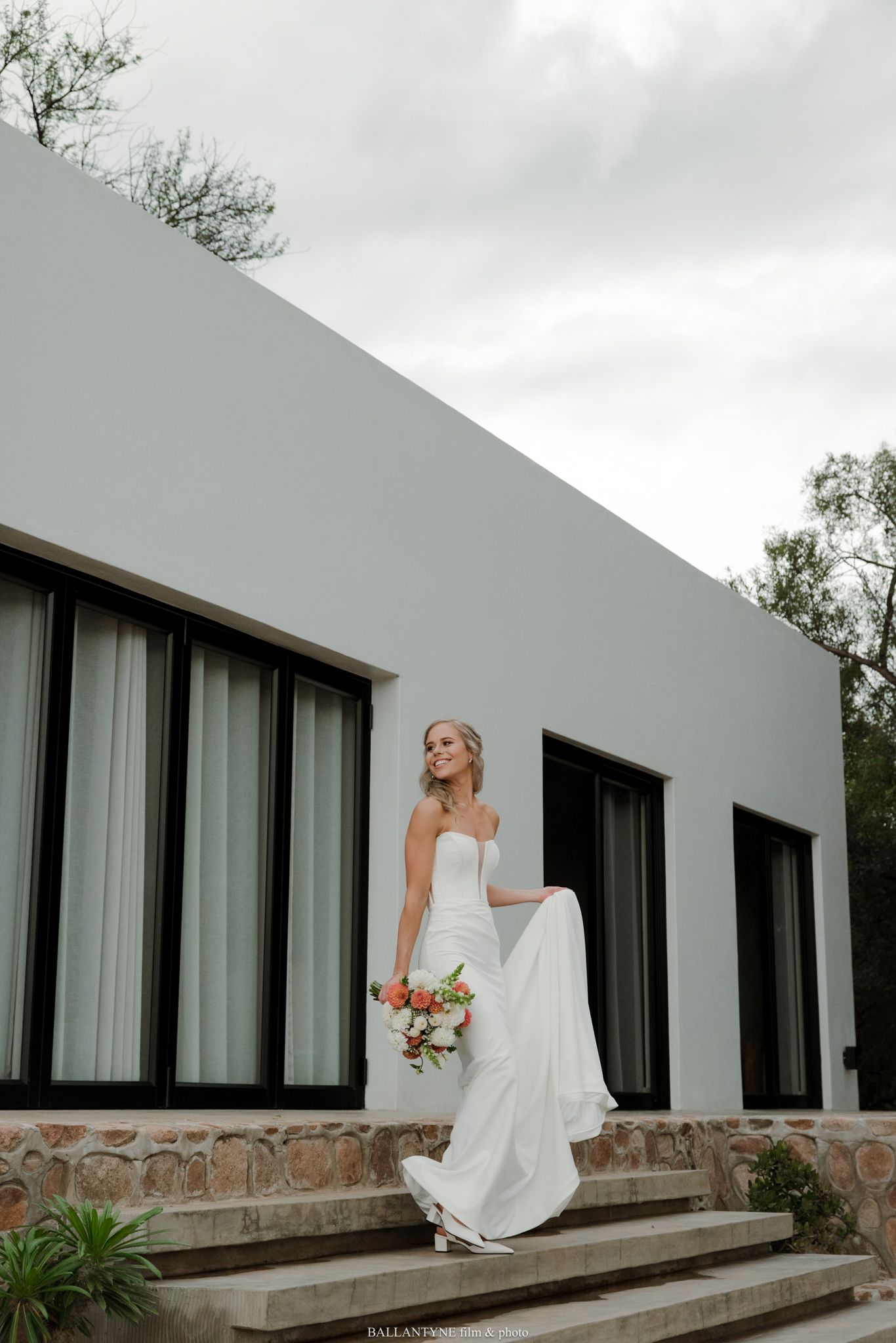 Bride in strapless white gown holding floral bouquet stands on stone steps outside modern gray building.