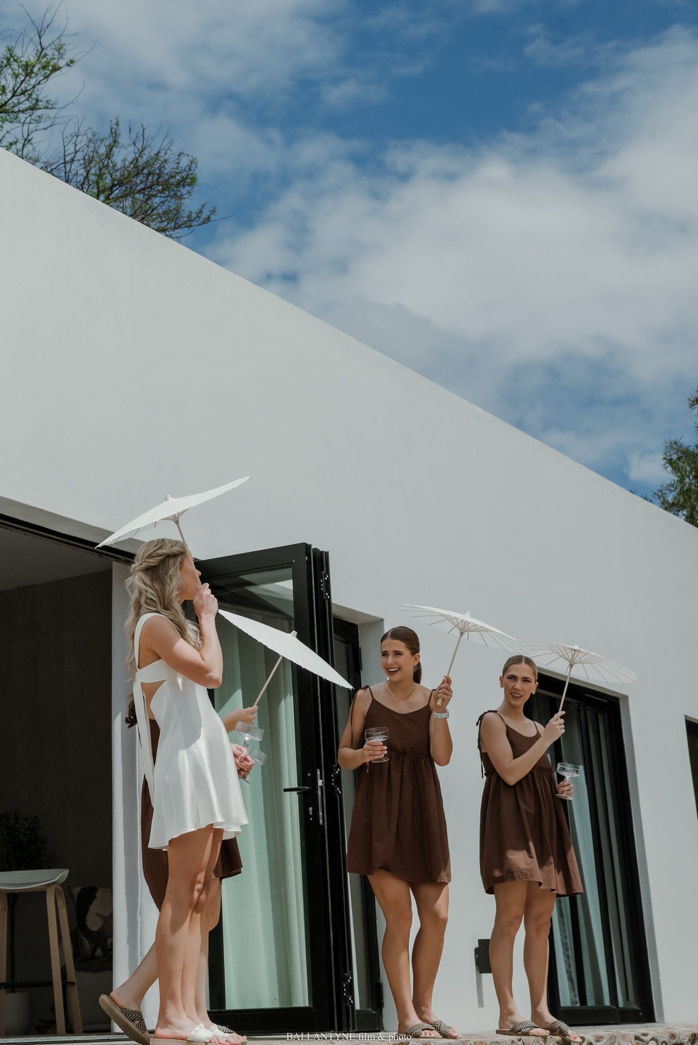 Bride in white dress and bridesmaids in brown dresses holding white parasols outside a modern white building.
