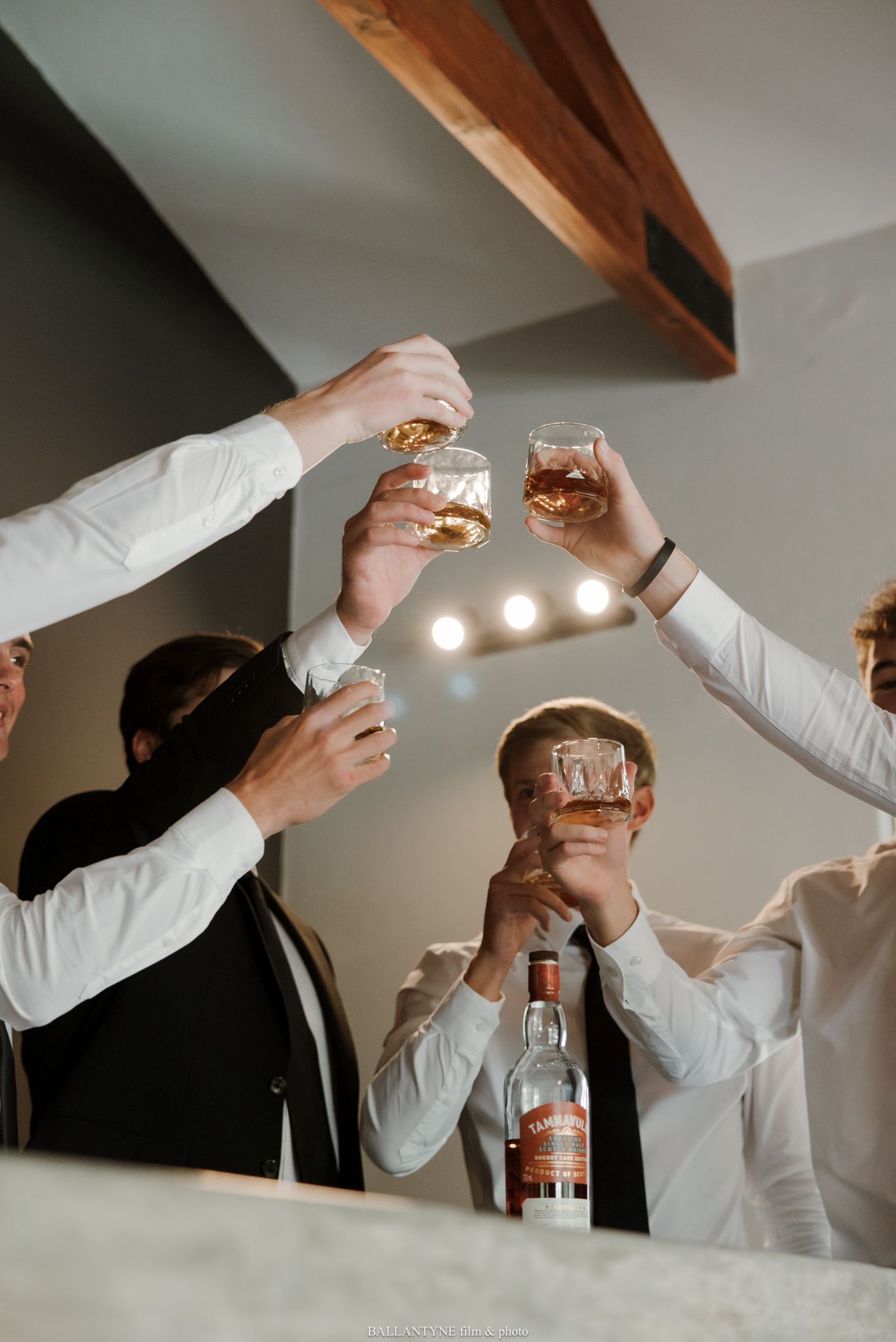 Men in dress shirts raising glasses of whiskey in a celebratory toast with a Tamnavulin bottle on the counter.