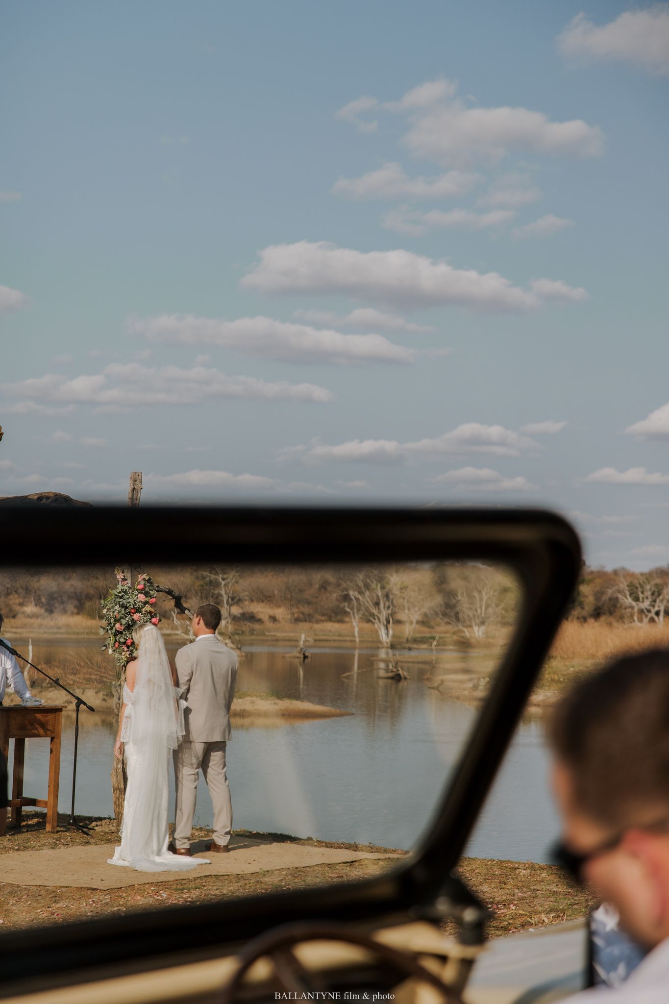 Wedding ceremony setup viewed through car window overlooking lake landscape.