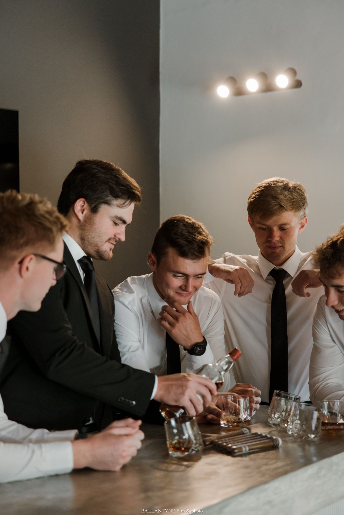 Five young men in dress shirts and ties gather around a bar counter examining whiskey glasses and cigars.