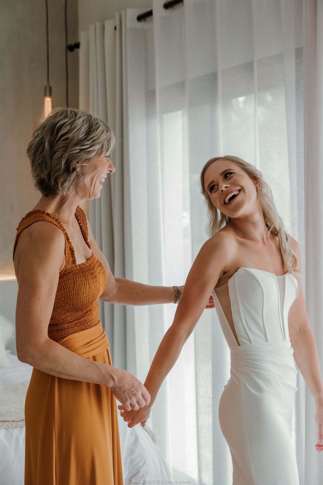 Bride in white corset gown laughing and holding hands with older woman in golden dress during wedding preparations.
