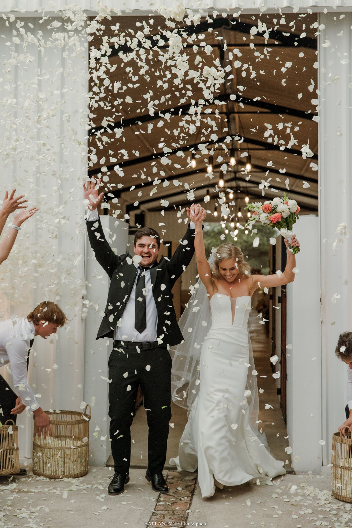 Bride and groom celebrate with confetti shower at wedding venue exit, arms raised in joy.