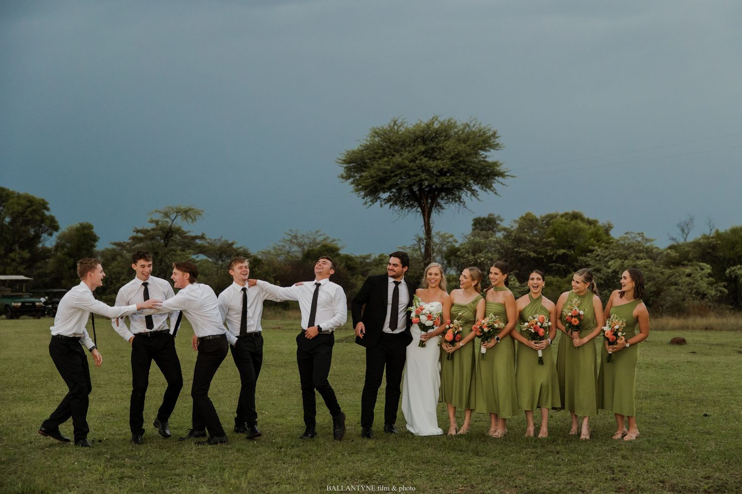 Wedding party poses playfully on green lawn with bride groom groomsmen and bridesmaids in sage dresses under stormy sky.