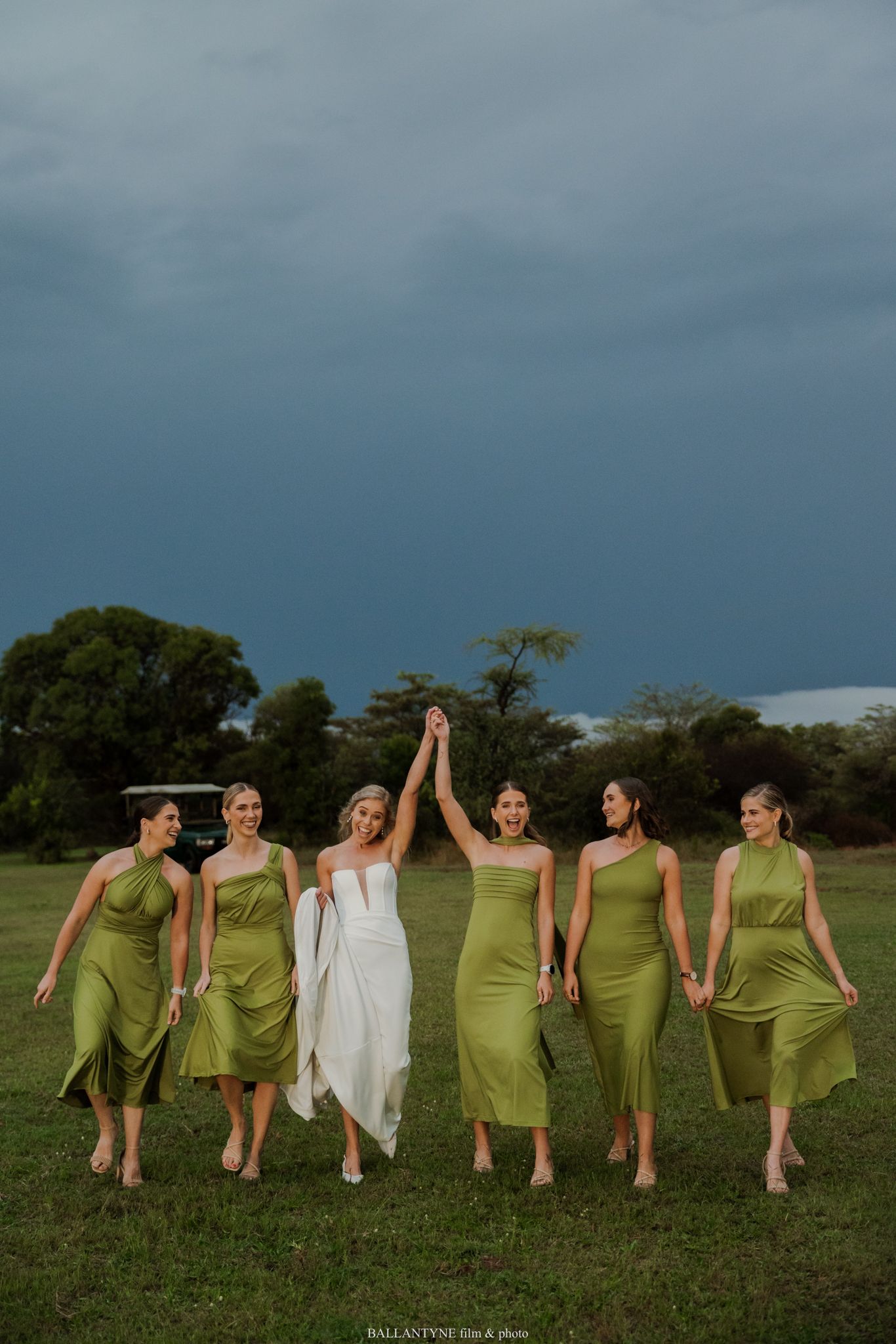 Bride in white gown raises hands with five bridesmaids in olive green satin dresses walking outdoors under dramatic stormy sky.