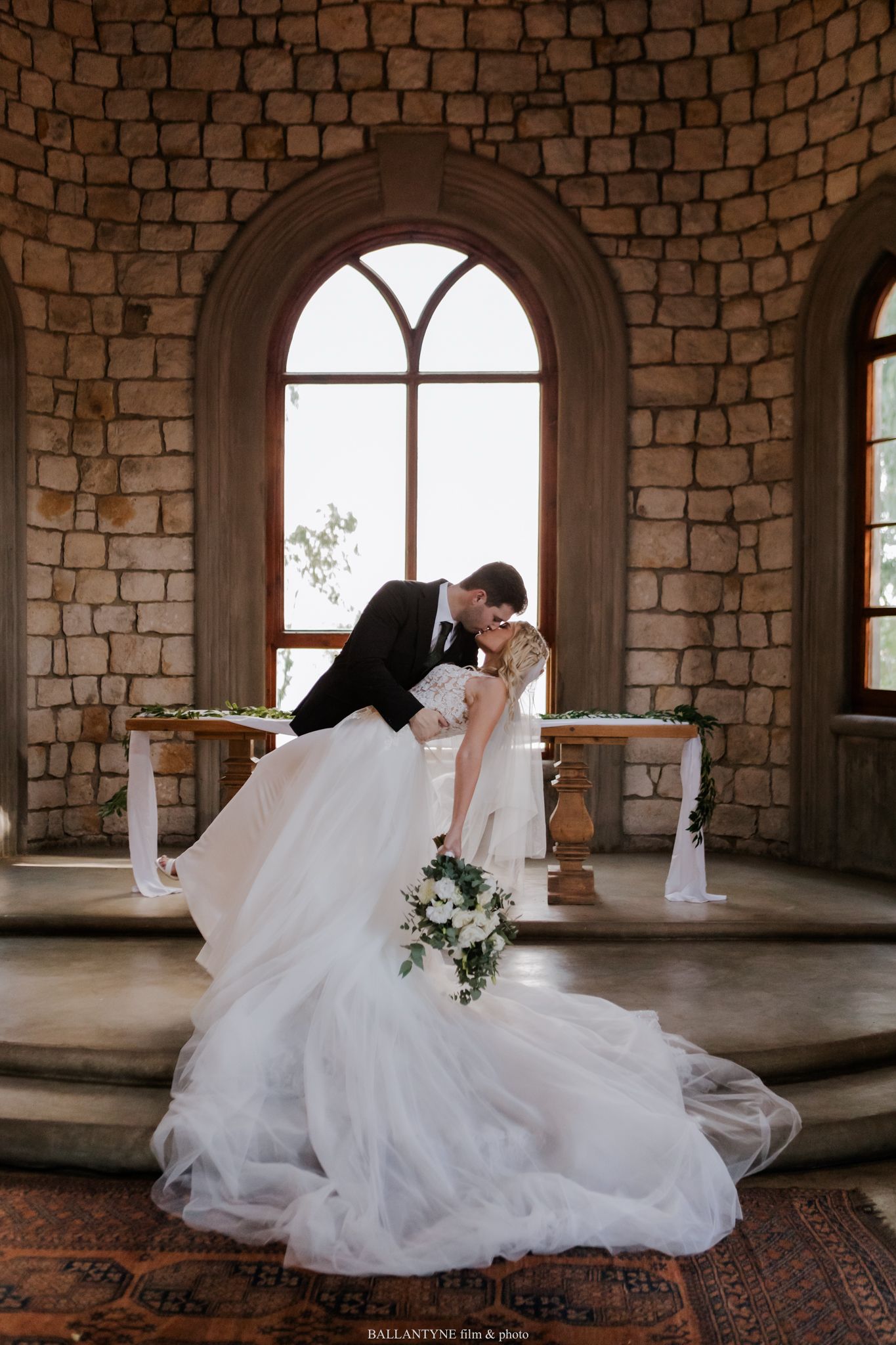 A romantic wedding photo in a stone castle room with an arched window and flowing white dress.