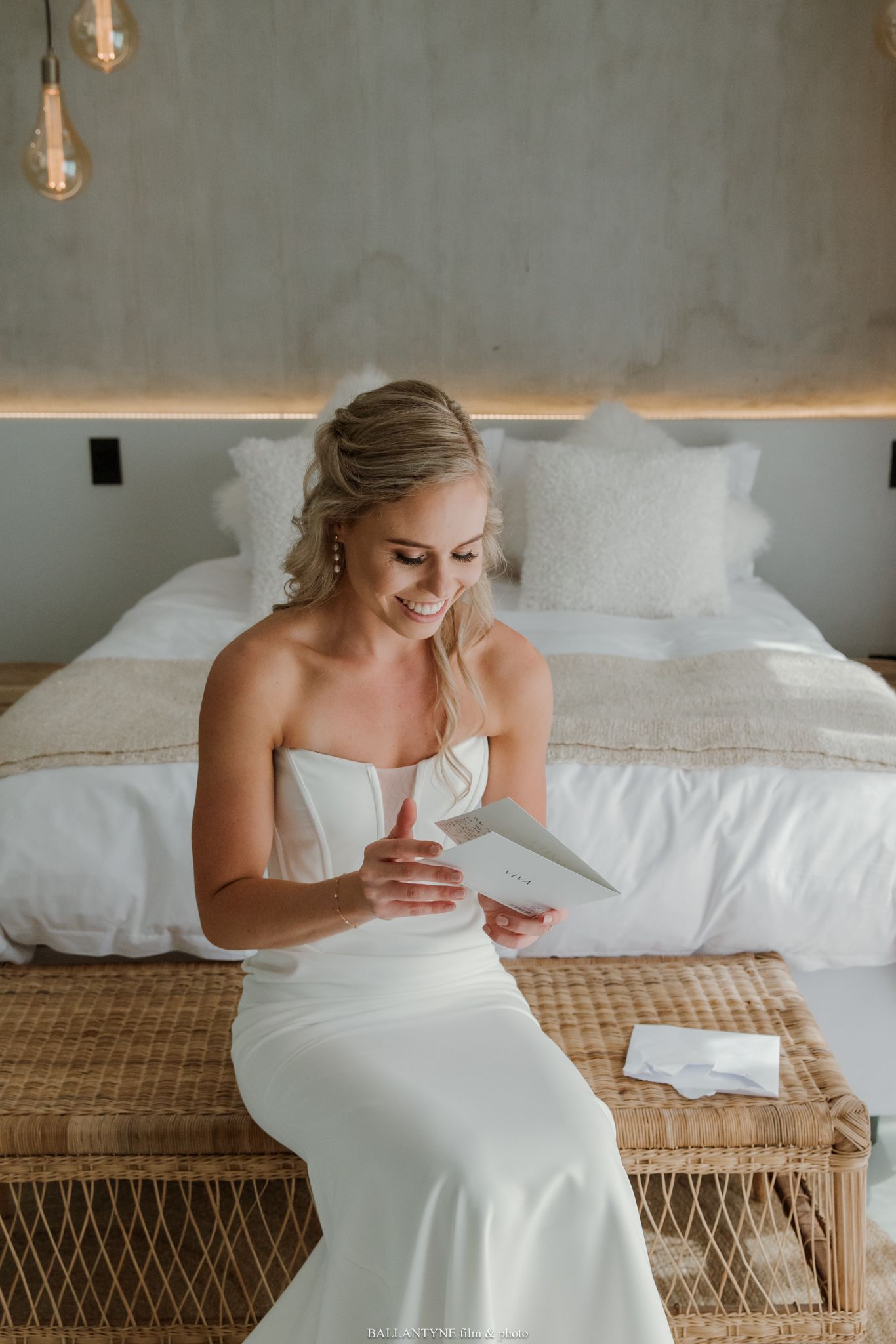 Smiling bride in white strapless gown reading a card while seated on a wicker bench in a modern bedroom.