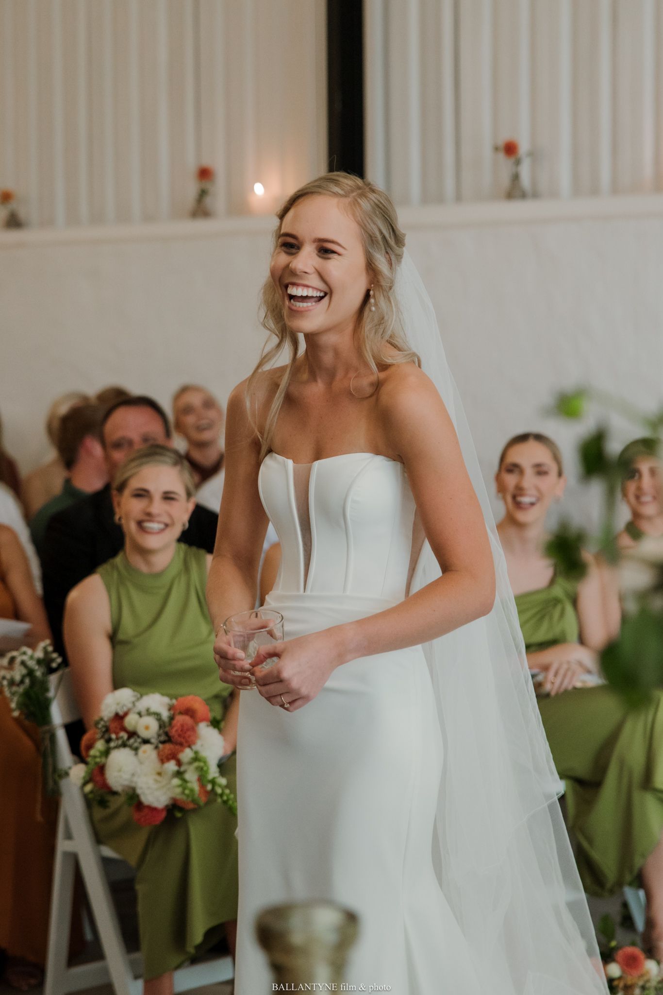 Bride in white strapless gown laughing joyfully at wedding ceremony with bridesmaids in green dresses seated behind her.