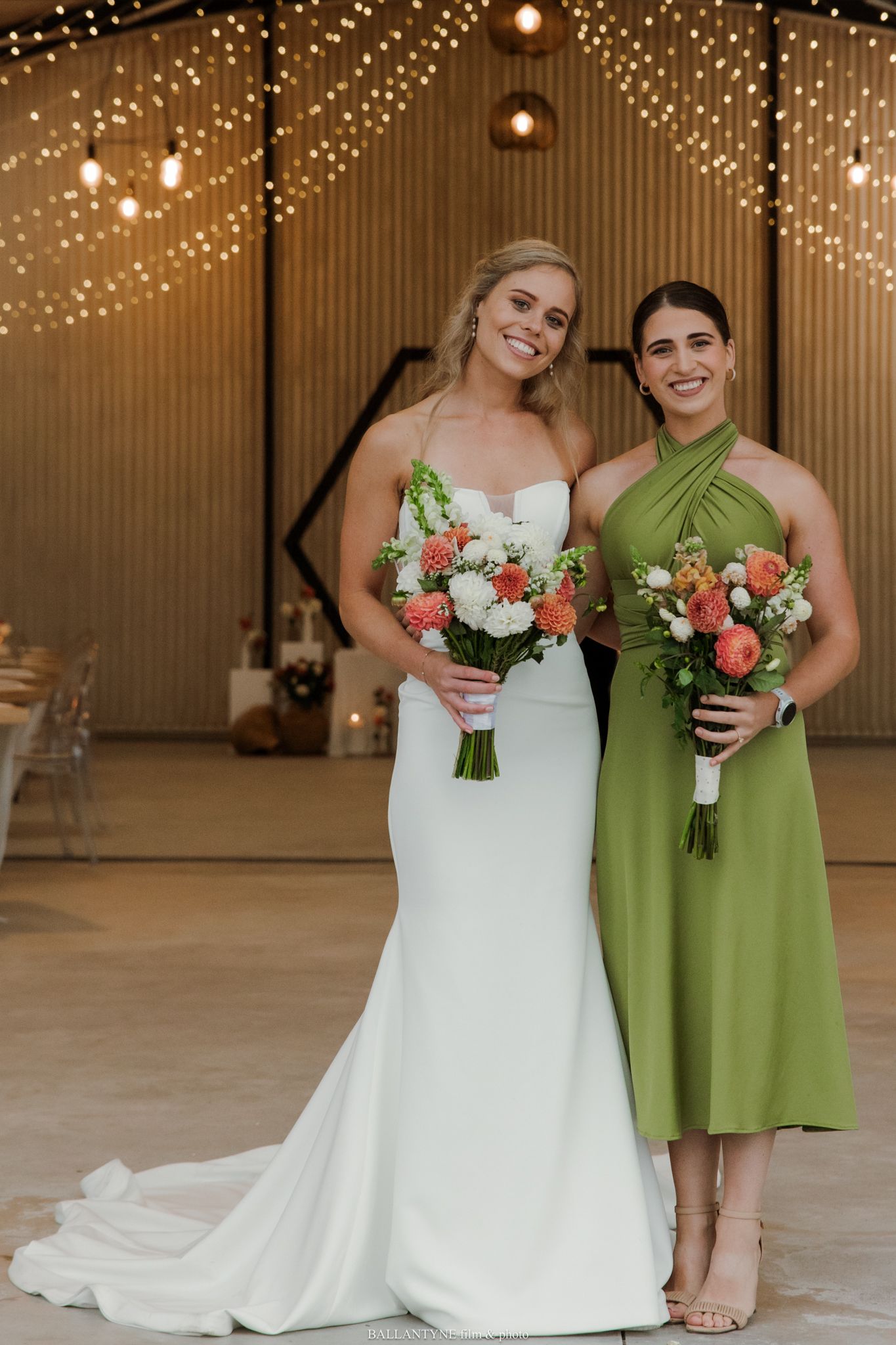 Bride in white gown and bridesmaid in green dress holding coral bouquets at a barn wedding venue.