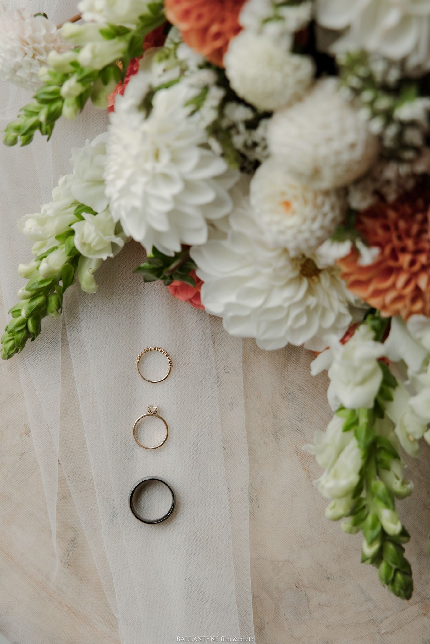Three wedding rings arranged on white veil fabric beside a bridal bouquet of white dahlias and coral flowers.