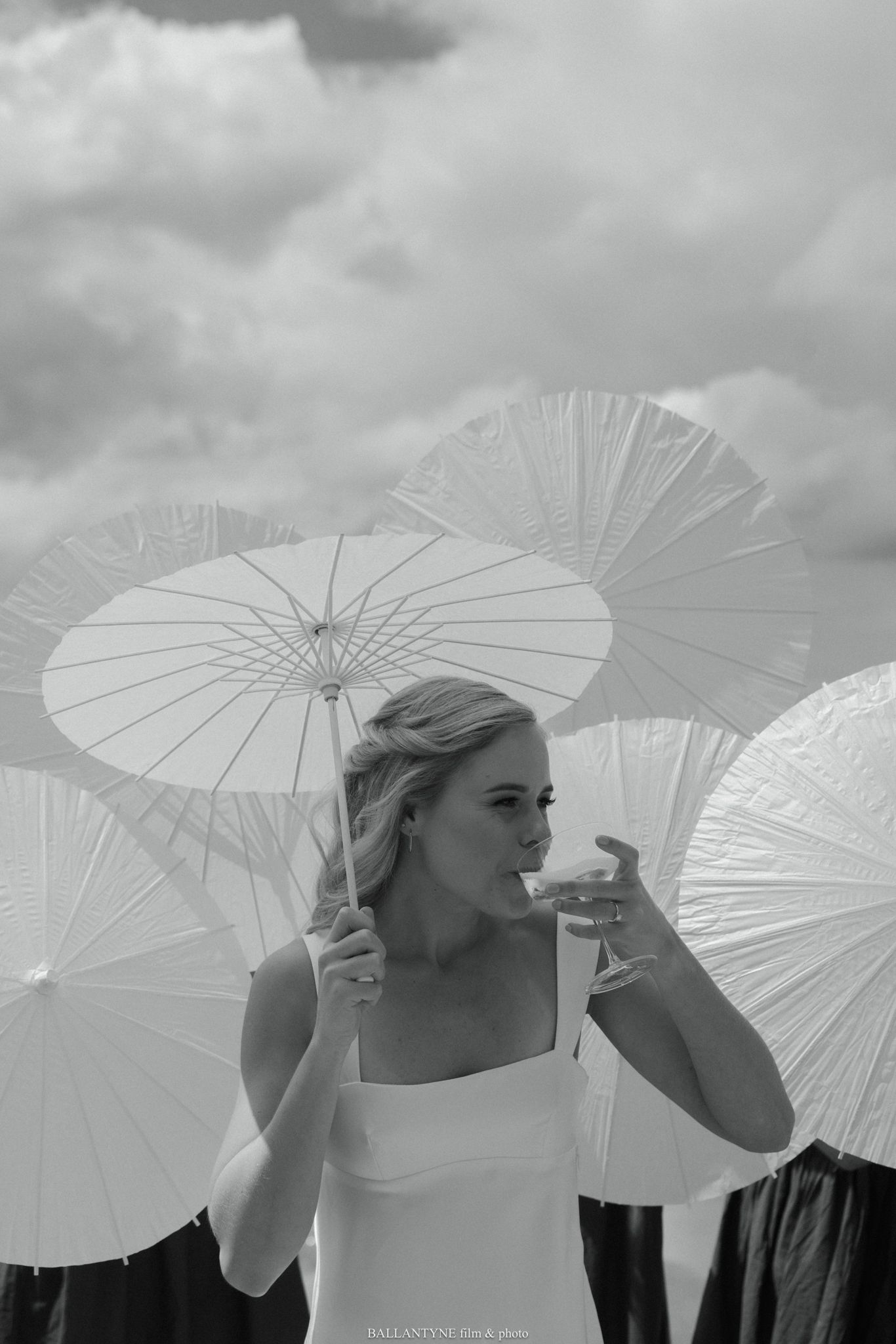 Black and white photo of a bride in white dress holding a champagne glass surrounded by paper parasols under cloudy sky.