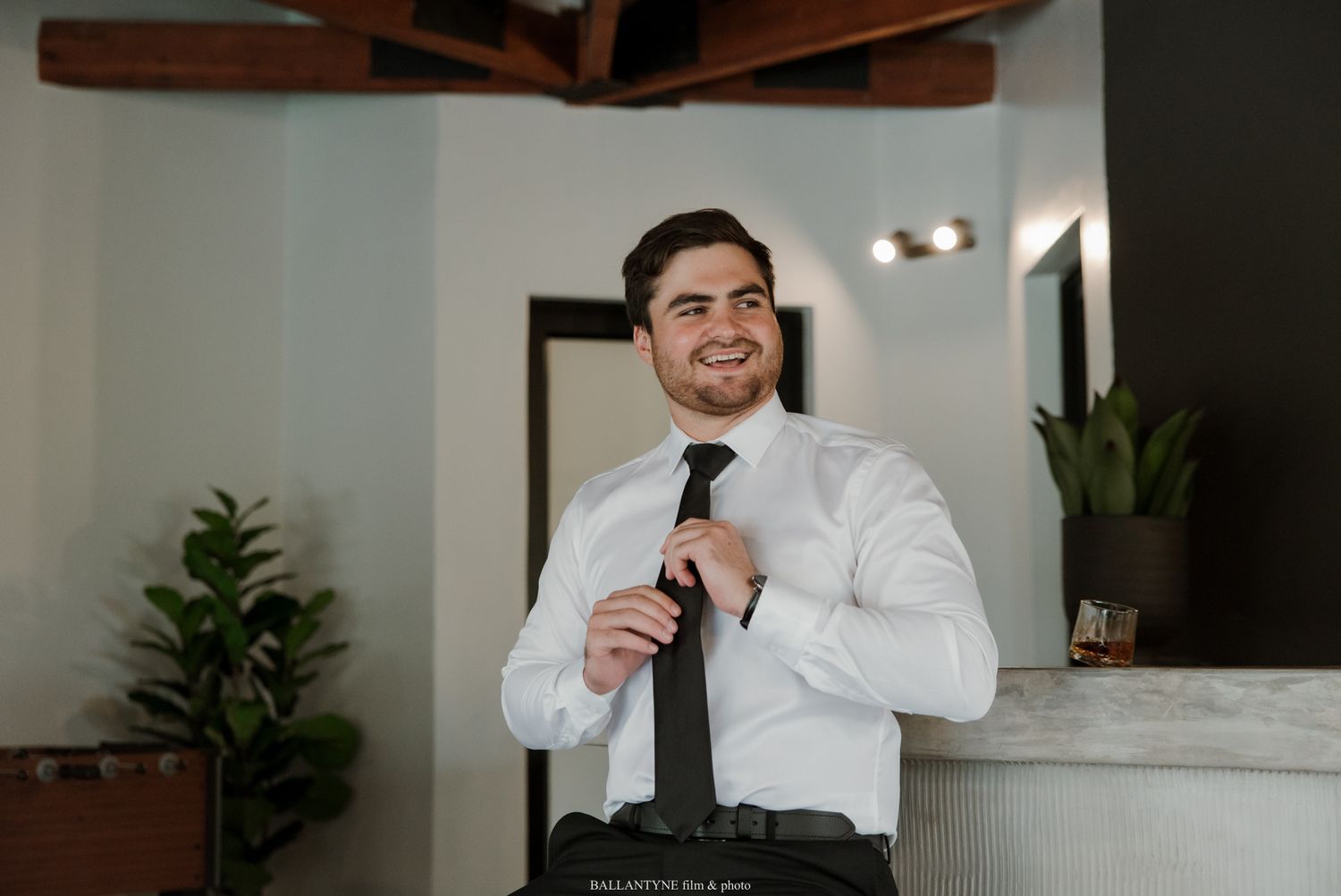 A smiling young man in a white dress shirt adjusting his black tie indoors.