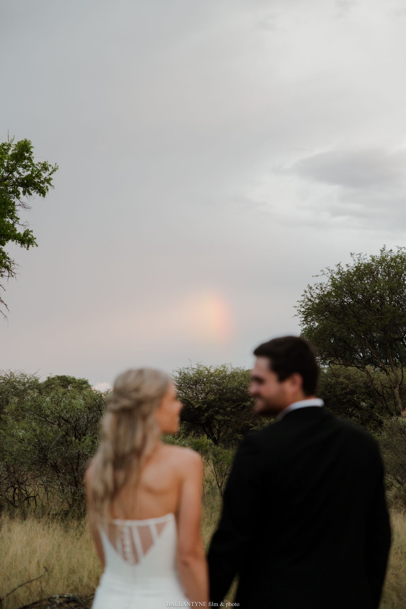 Bride and groom holding hands facing a glowing sunset sky over lush bushveld landscape on their wedding day.