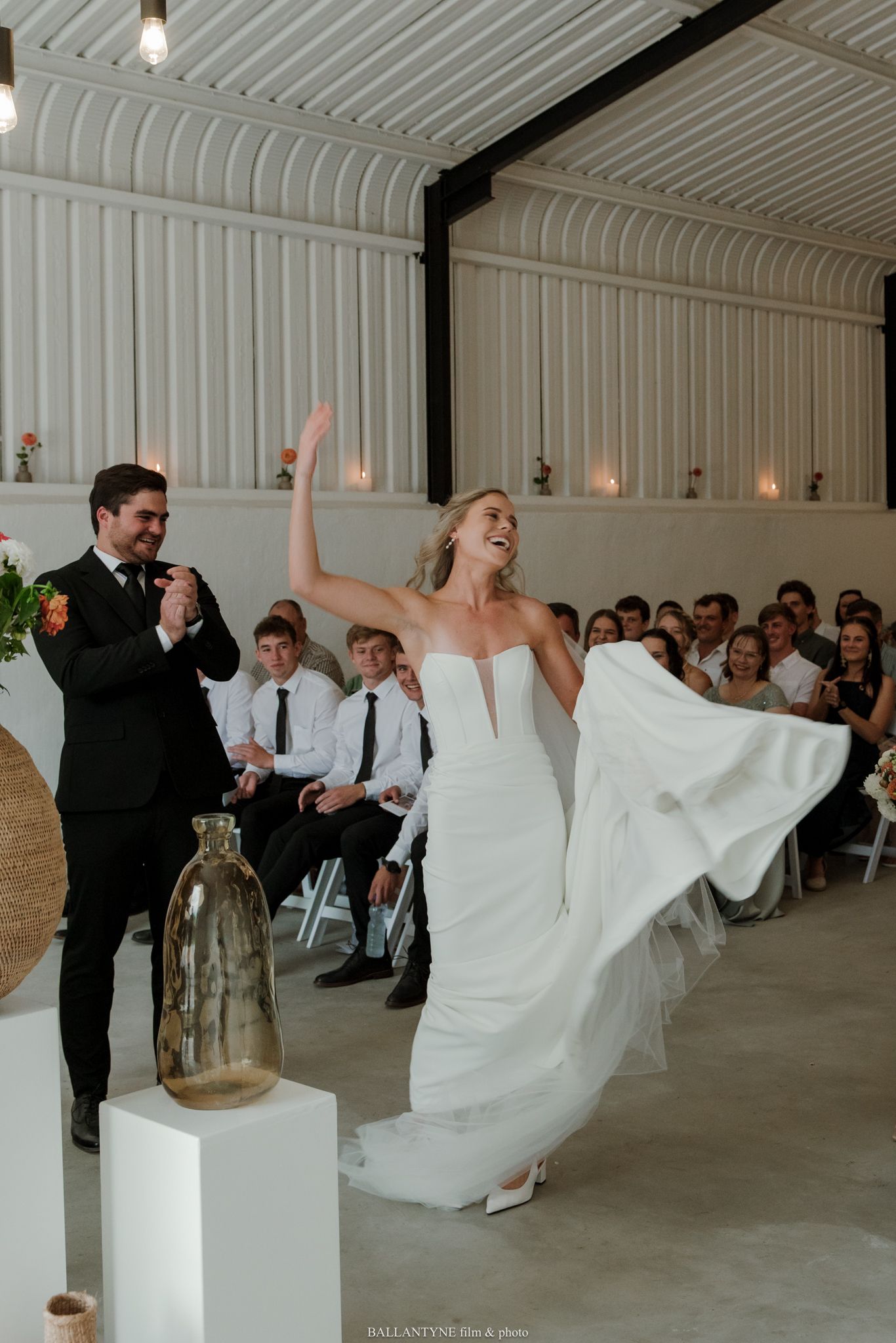 Joyful bride in white gown twirling at wedding reception while groom applauds, guests seated in background.