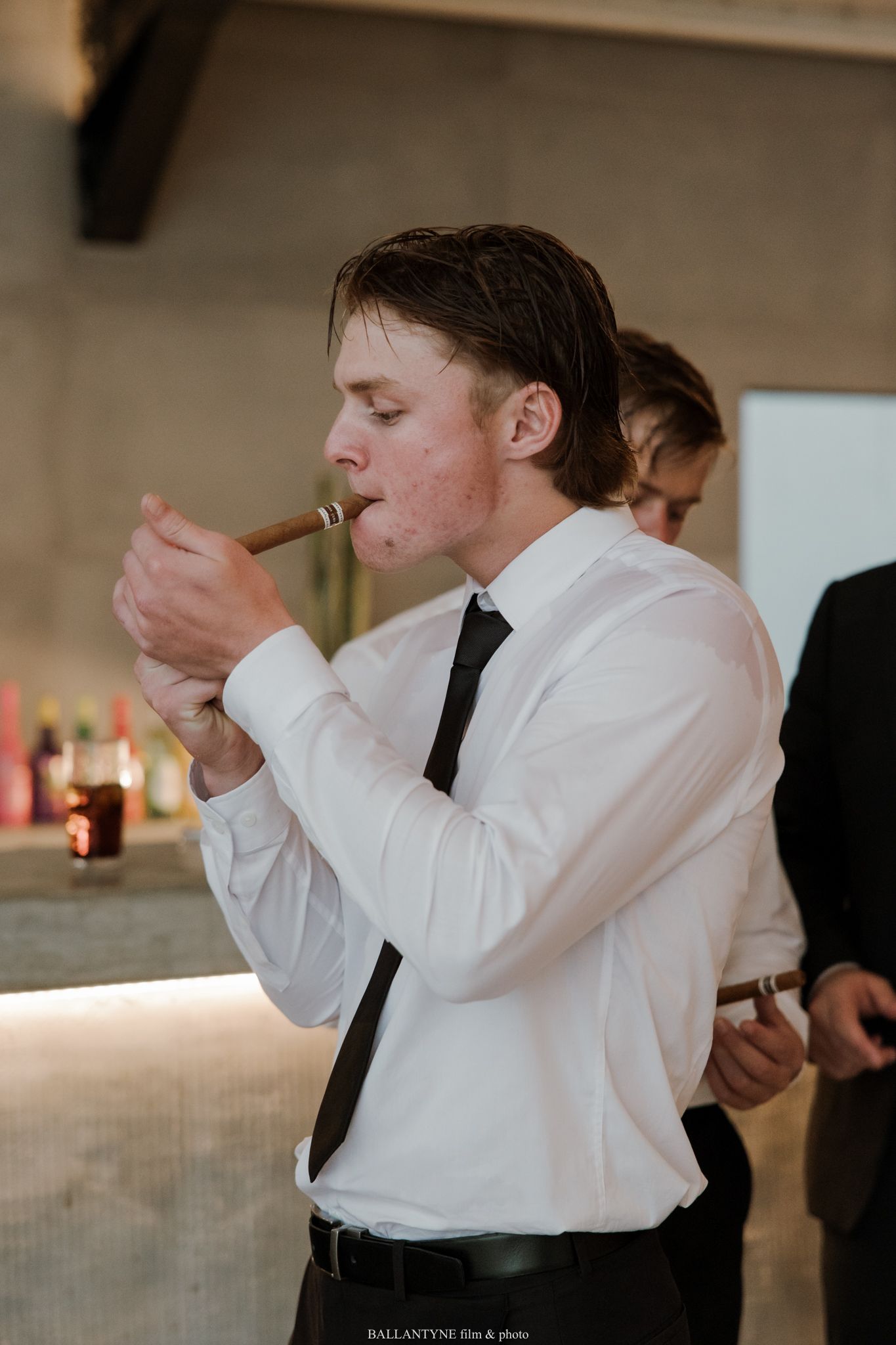 Young man in white shirt and black tie lighting a cigar at a formal event with a bar in the background.