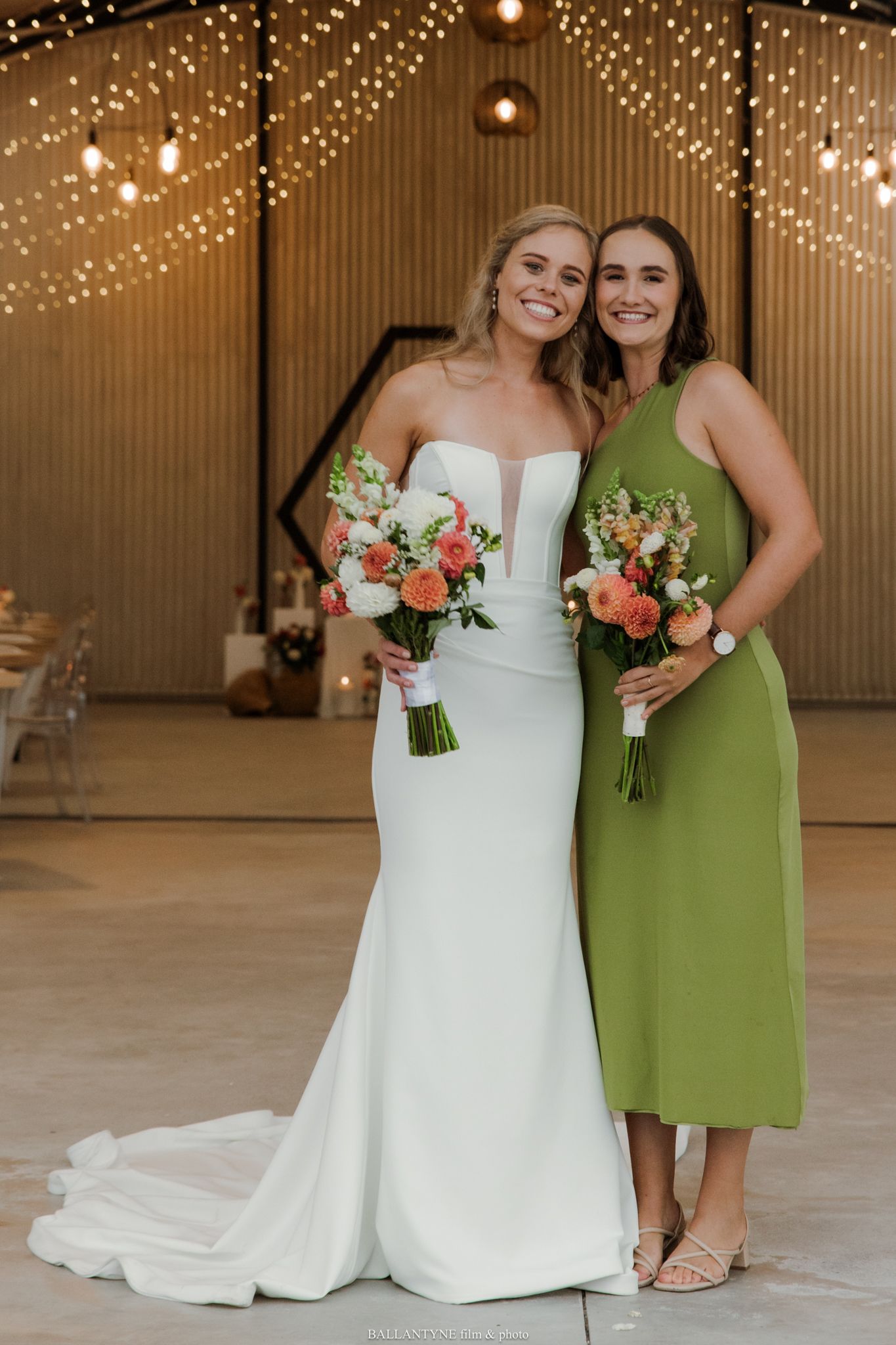Bride in white gown and bridesmaid in green dress holding coral bouquets at elegant barn wedding venue.