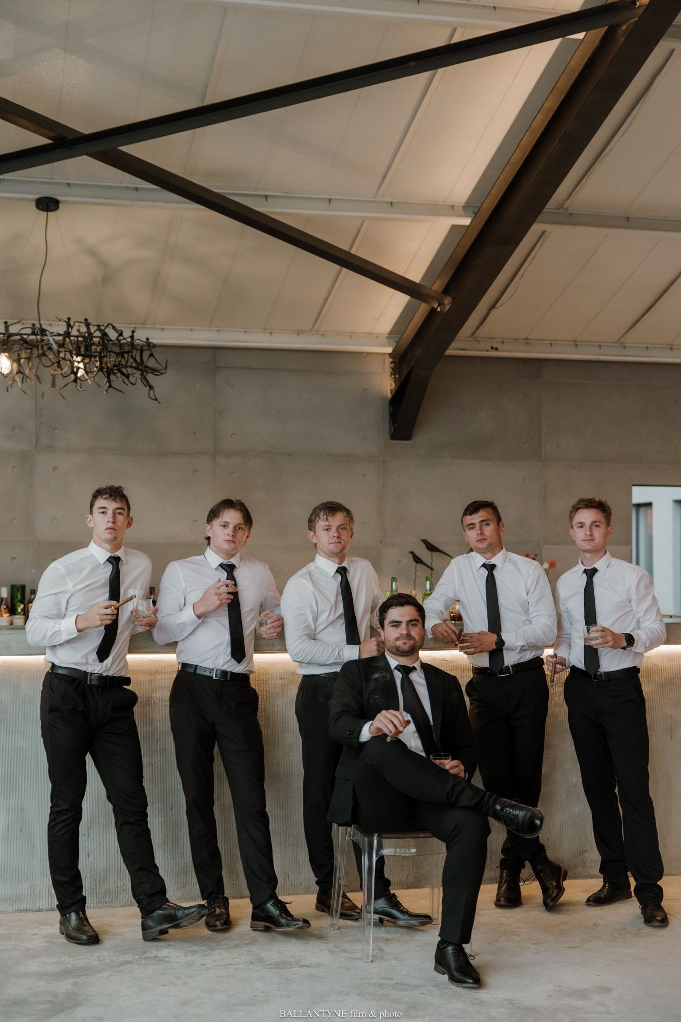 Groom in black suit seated confidently with five groomsmen in white shirts and black ties posing behind him indoors.