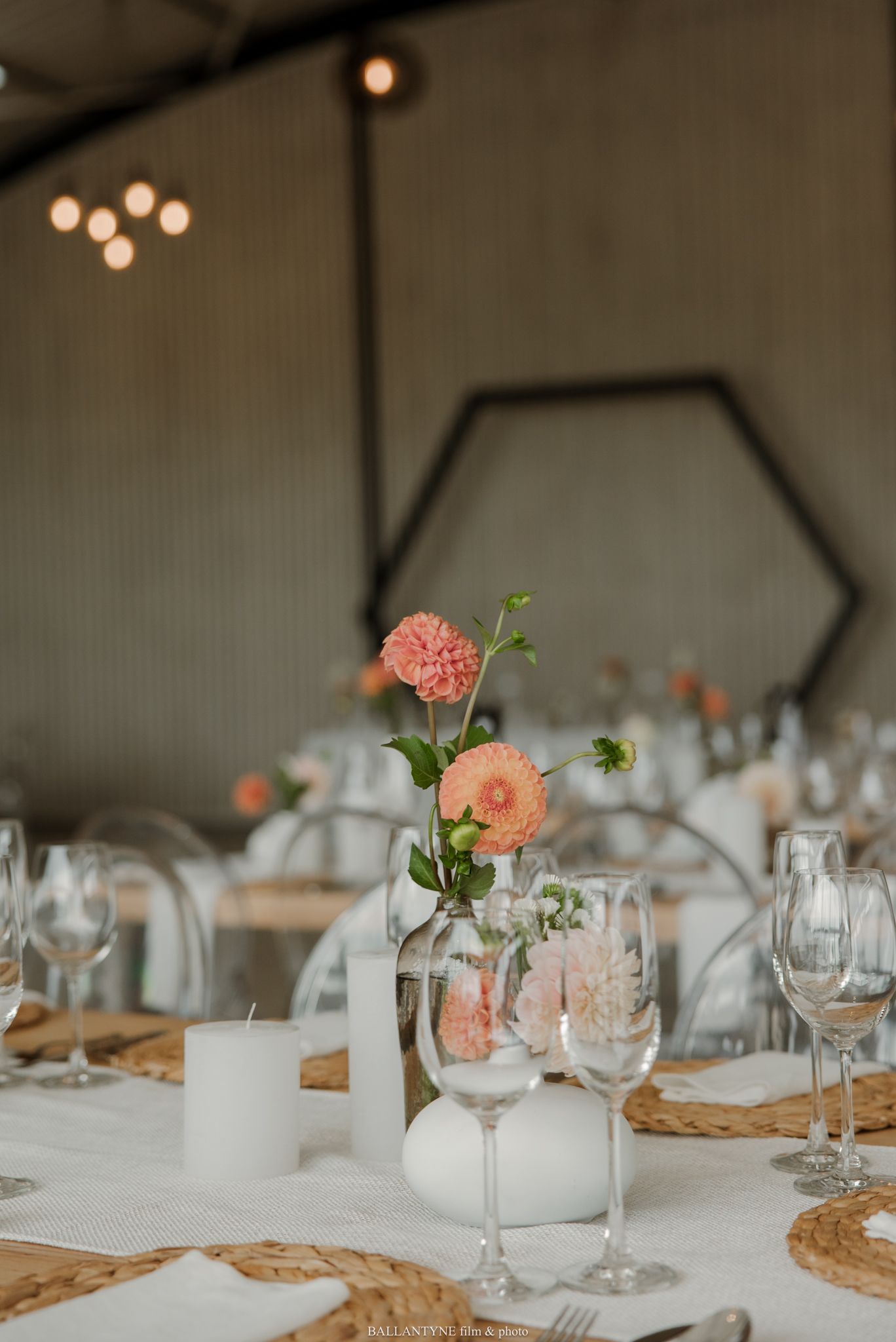 Elegant wedding table setting with peach flowers, white candles, wine glasses, and woven placemats in a softly lit venue.