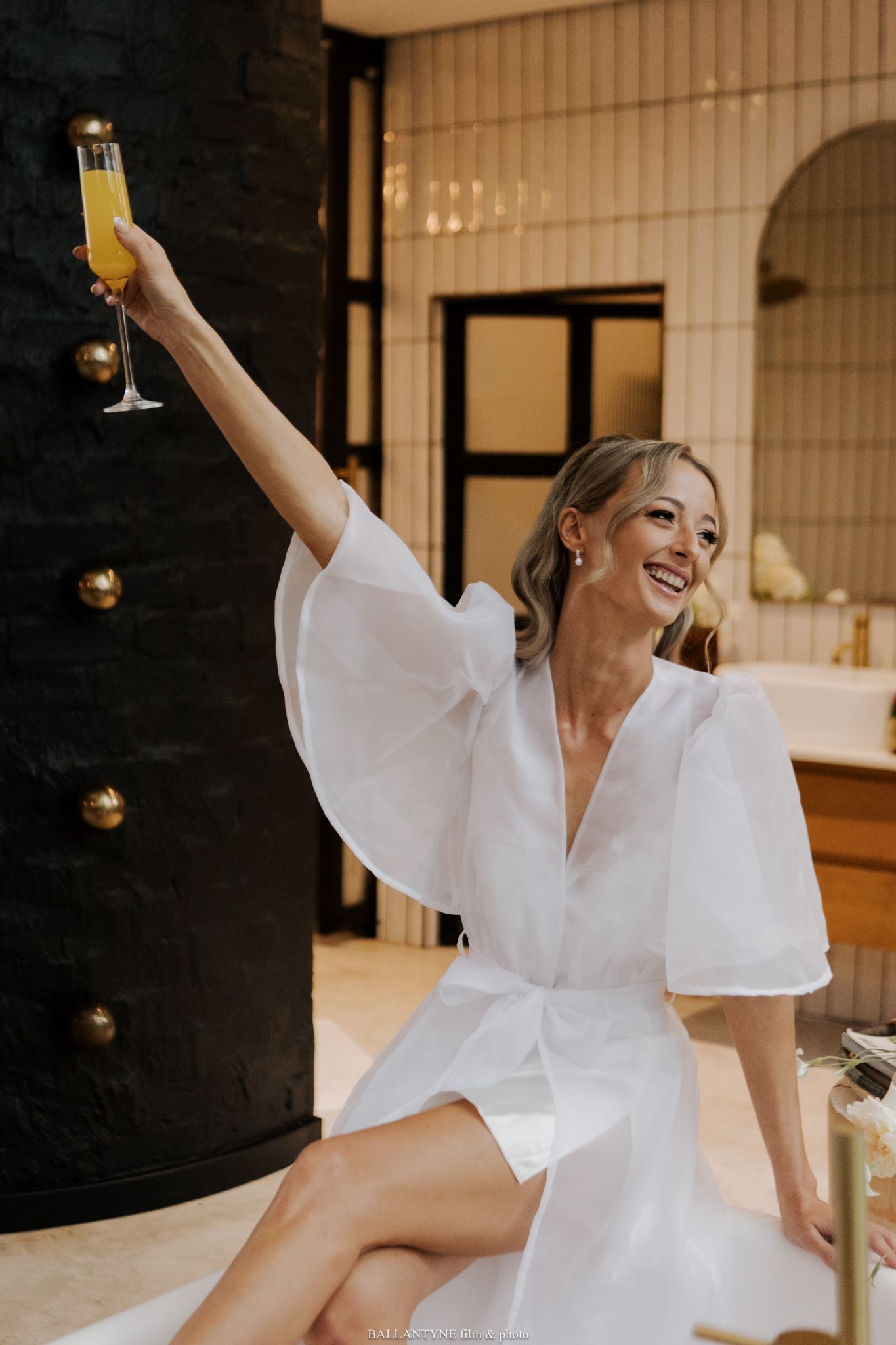 Bride in white robe celebrating with champagne in an indoor setting.