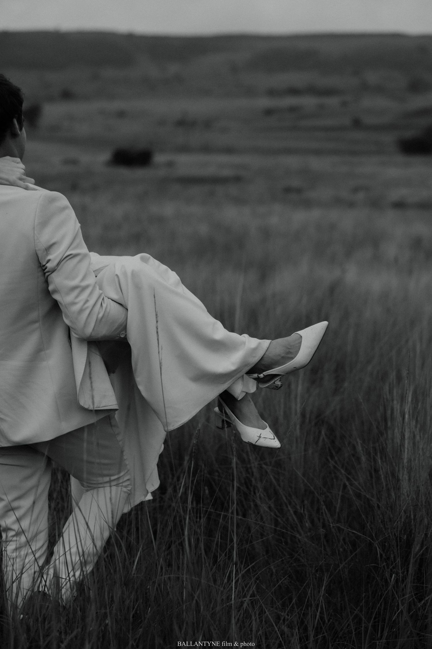 Black and white photo of a romantic moment in a field with flowing wedding dress and formal attire.