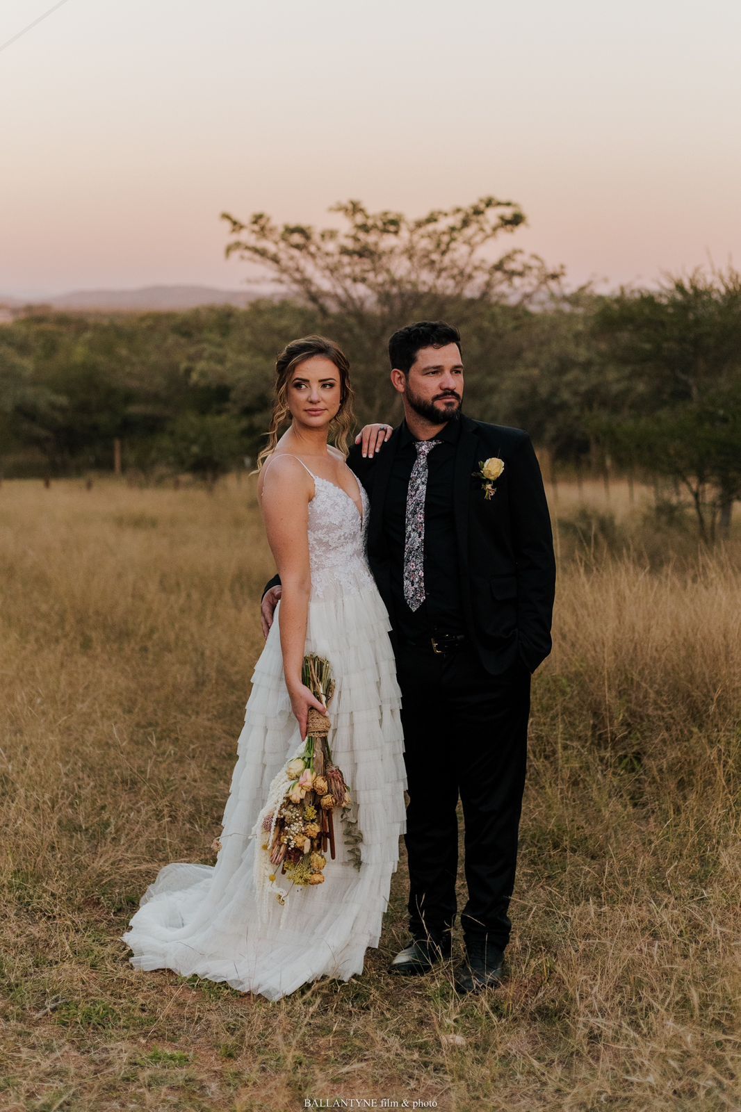 A couple in wedding attire poses together in a rustic field at sunset.