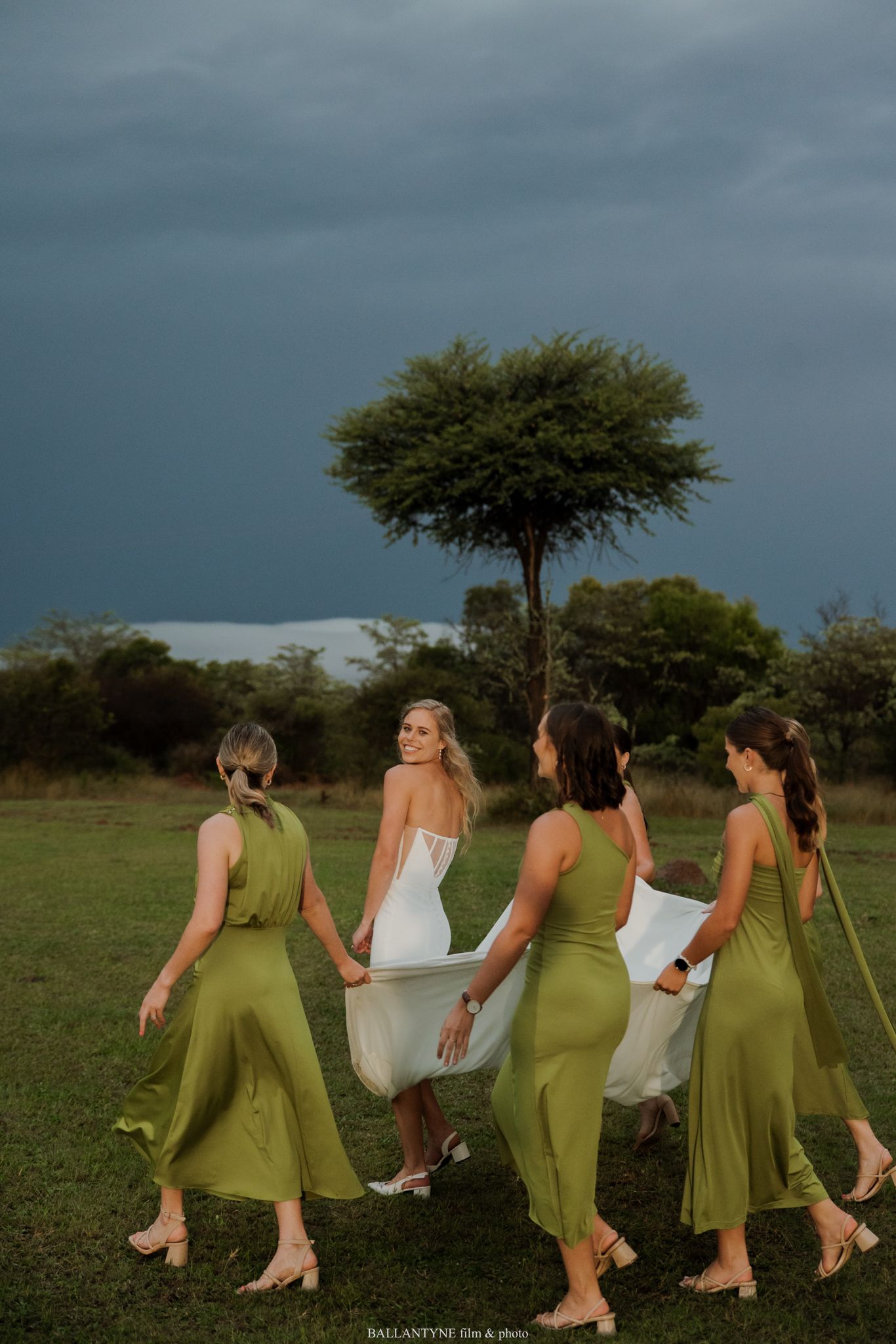 Bride in white gown walks with three bridesmaids in olive green dresses across a lush field under a dramatic stormy sky.