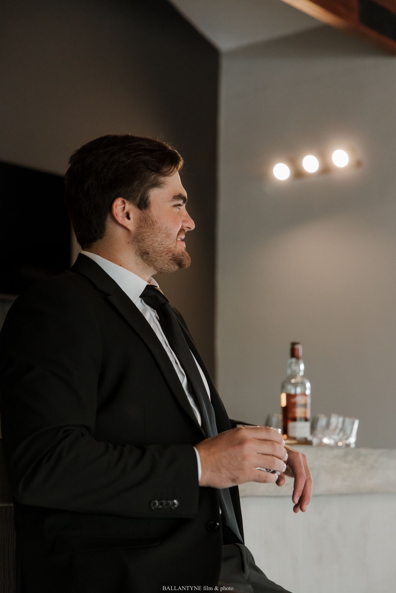 Groom in black suit holding a drink, smiling at a bar with whiskey bottle and glasses in background.