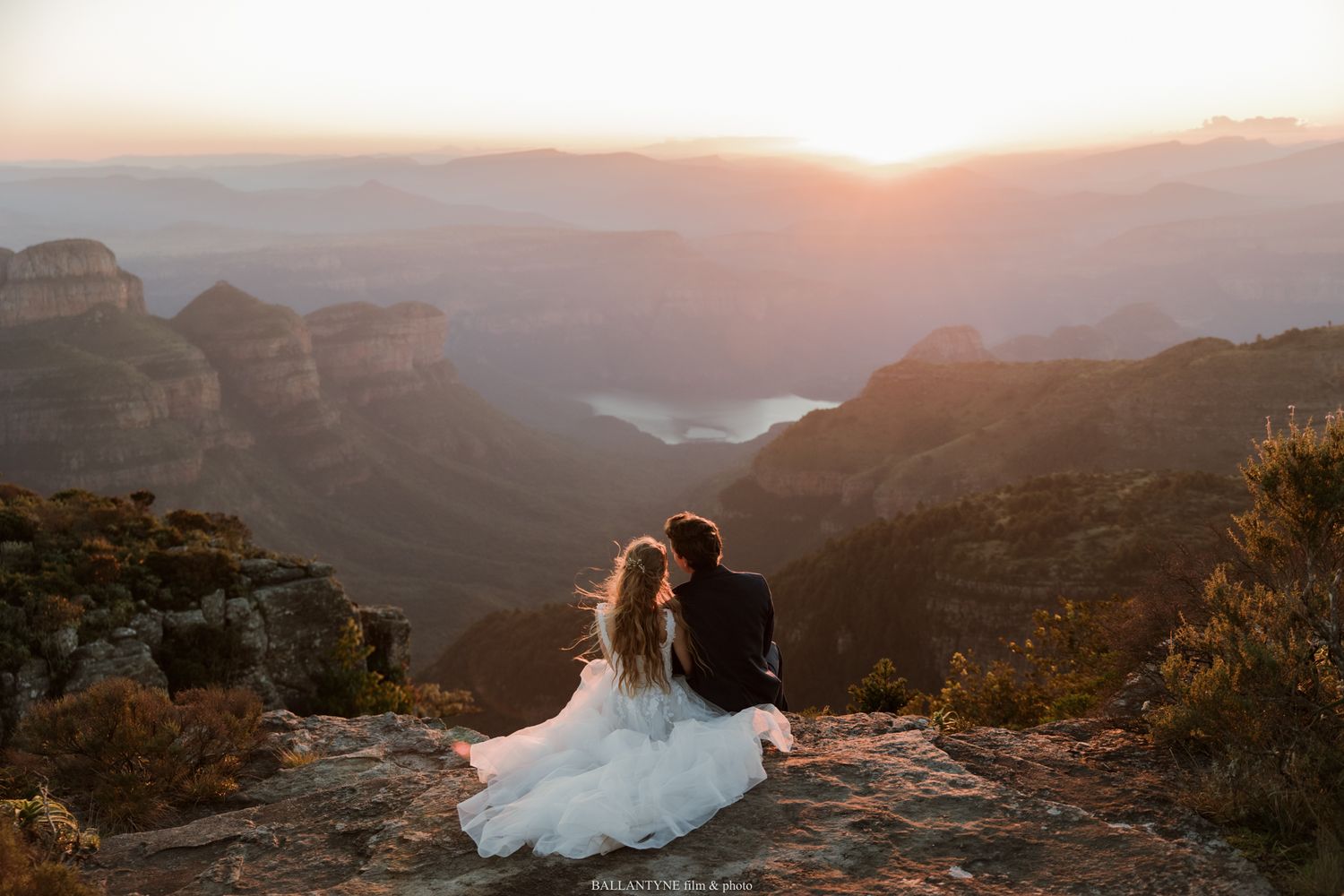 Bride and groom sit together on a rocky cliff edge at sunset overlooking a stunning canyon landscape.