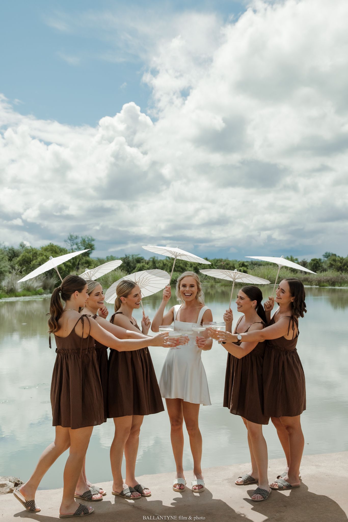 Bride in white dress with bridesmaids in brown dresses holding white parasols, toasting by a lake under cloudy skies.