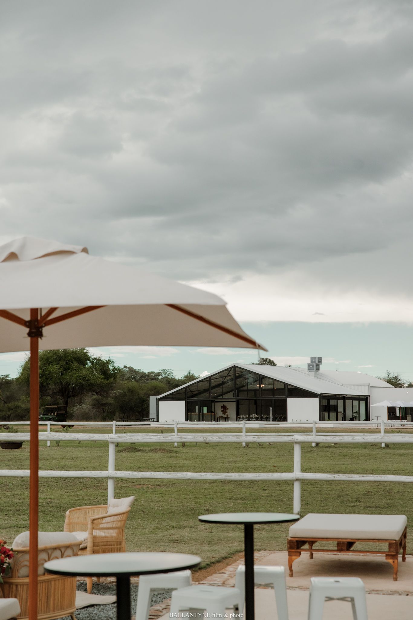 Outdoor patio with beige umbrella and rattan chairs overlooking a white fenced paddock and modern glass barn venue under cloudy skies.