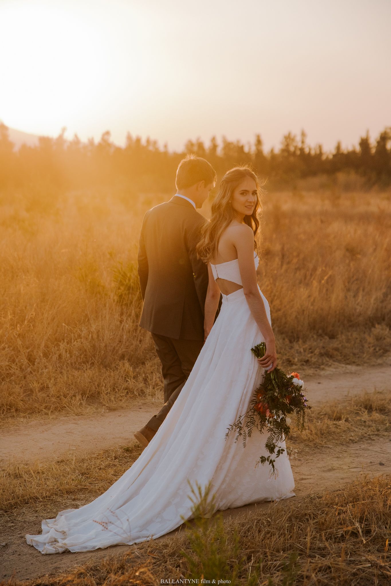 Bride in flowing white dress walking through golden field at sunset.