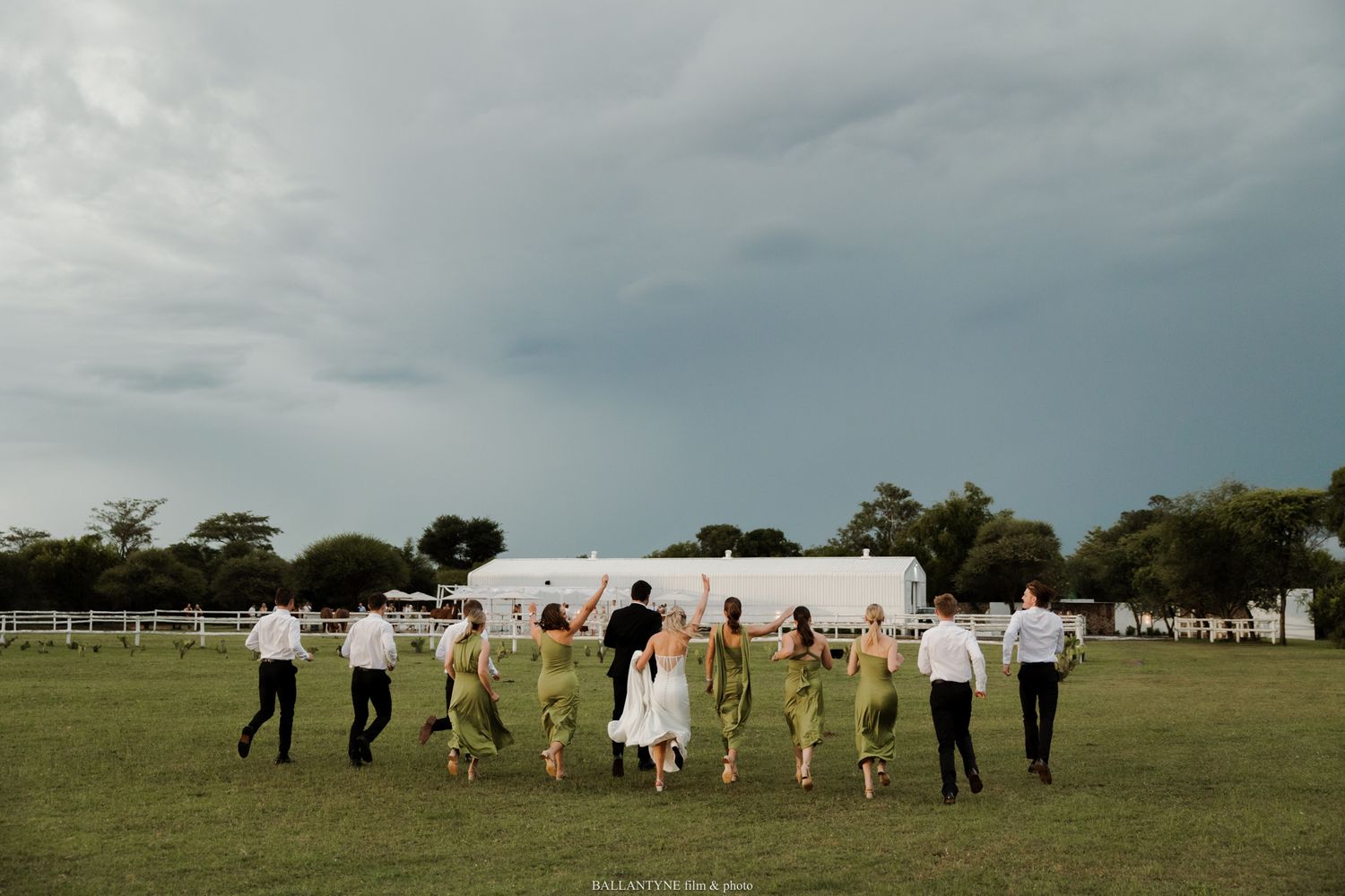 Wedding party in green dresses and black suits running across a green field under a dramatic stormy sky.