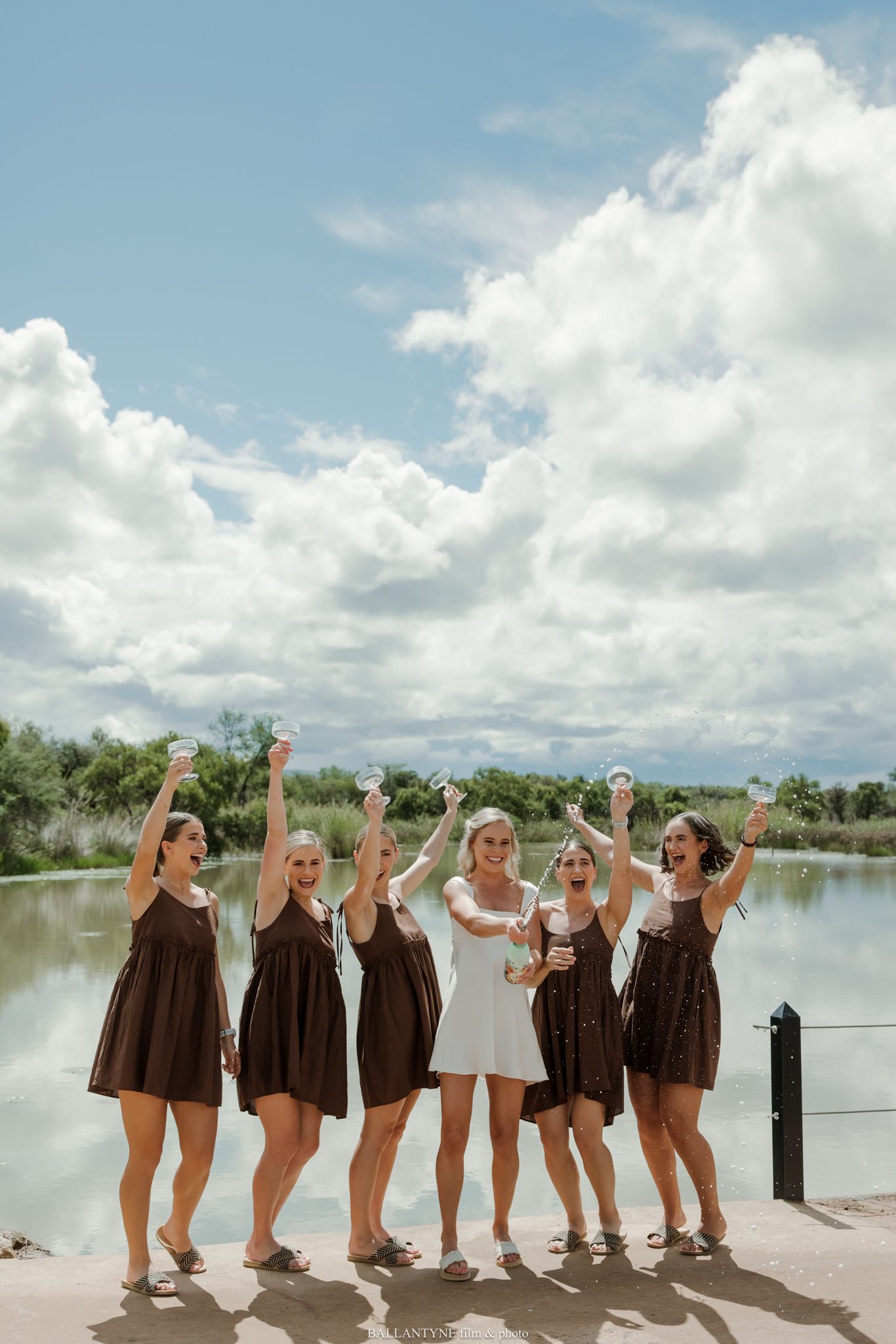 Bride in white dress celebrates with six bridesmaids in brown dresses by a lake under a cloudy sky.