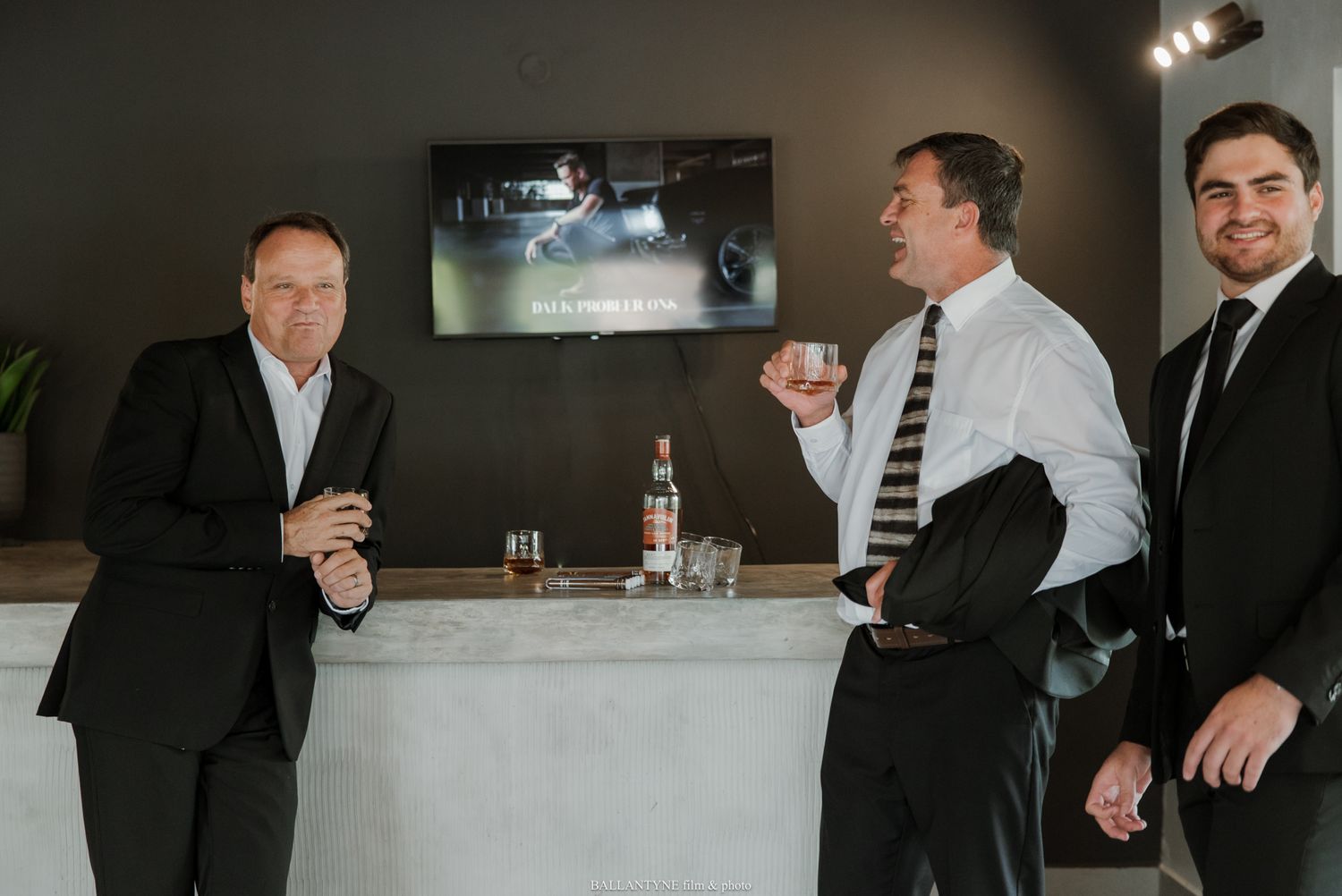 Three men in suits socializing at a modern bar counter, holding drinks and smiling in an elegant indoor setting.