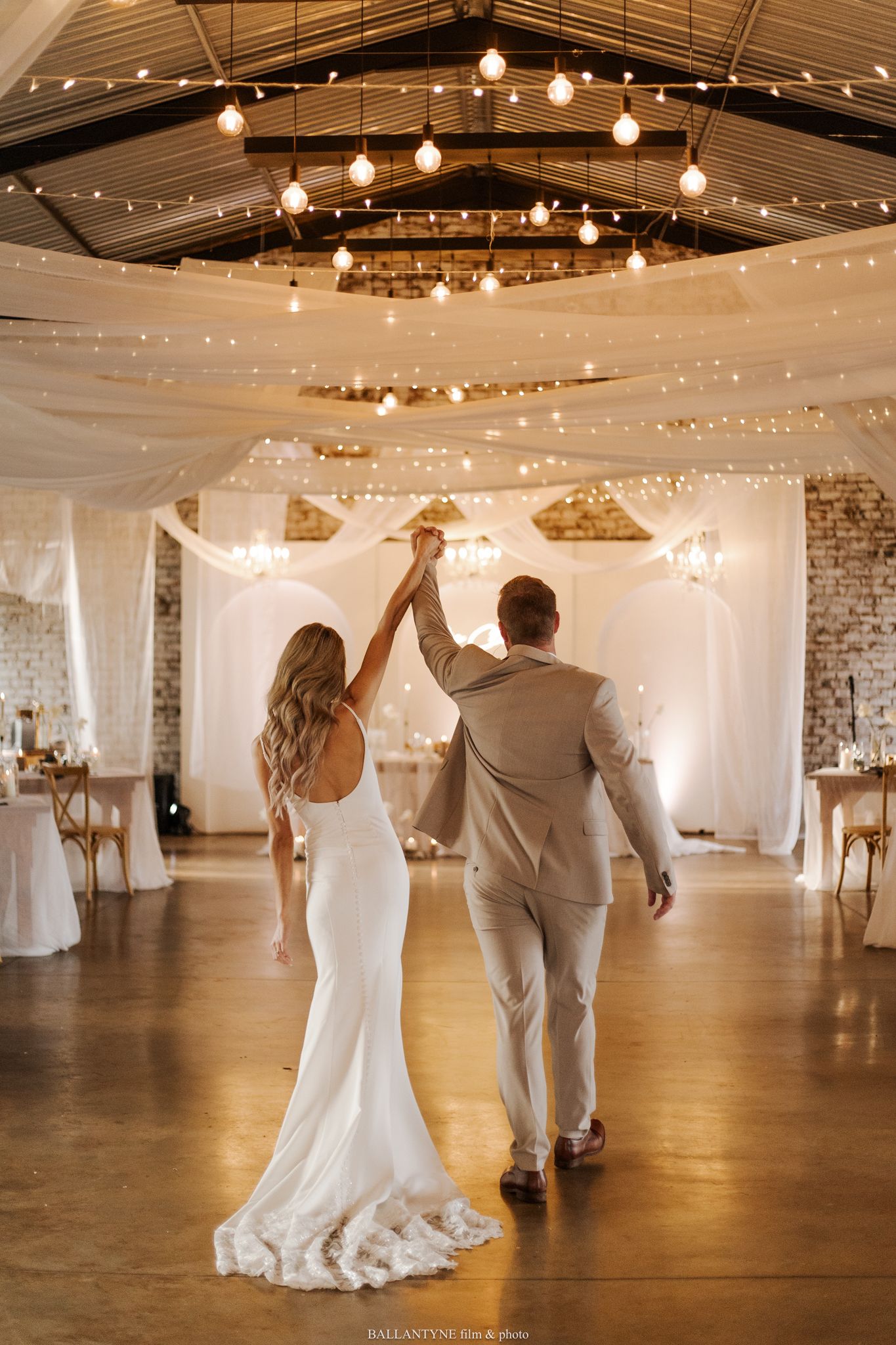 A couple dances in a beautifully decorated reception venue with string lights and white drapes.
